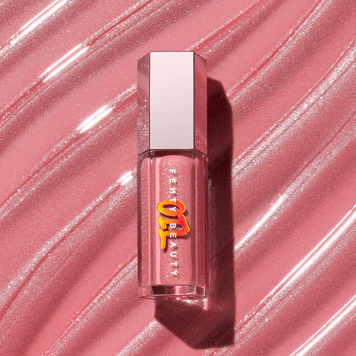 Fenty Beauty lipgloss in een heldere, zeshoekige verpakking met een zilveren dop, liggend op een glanzende, roze achtergrond.