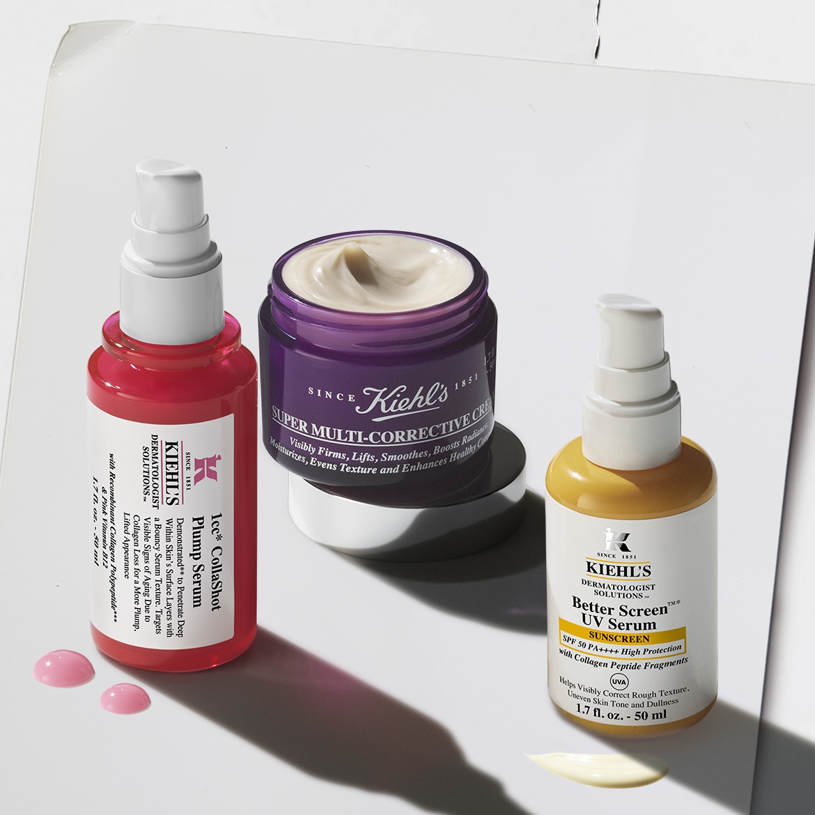 Drie Kiehl's producten staan op een witte ondergrond: een roze flesje 1cc CollaShot Plump Serum, een paarse pot Super Multi-Corrective Cream en een geel flesje Better Screen UV Serum, met van elk product een beetje crème ernaast.