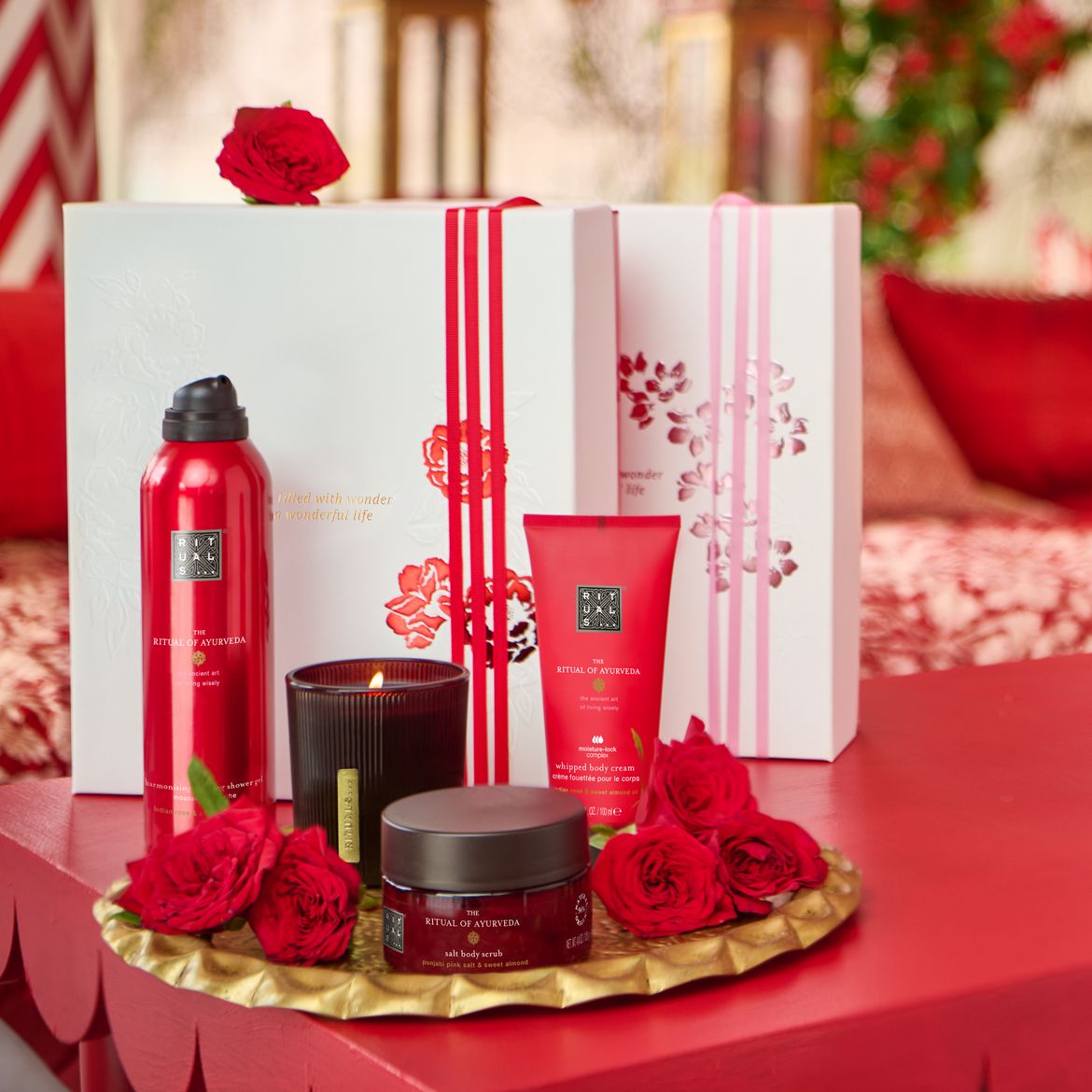 Ontdek de Ritual of Ayurveda-collectie, met een rode douchegel, bodycrème, scrub en geurkaars, gepresenteerd op een gouden schaal met rode rozen, naast een witte geschenkdoos met rode en roze linten.