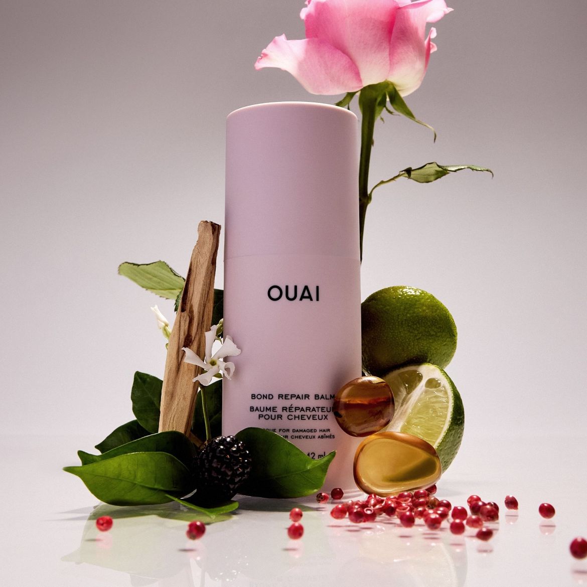 De Ouai Bond Repair Balm, een lichtroze tube van 42 ml, staat rechtop tussen verse ingrediënten zoals een roze roos, limoenen, bessen en houten takjes, wat de natuurlijke samenstelling van het product benadrukt.