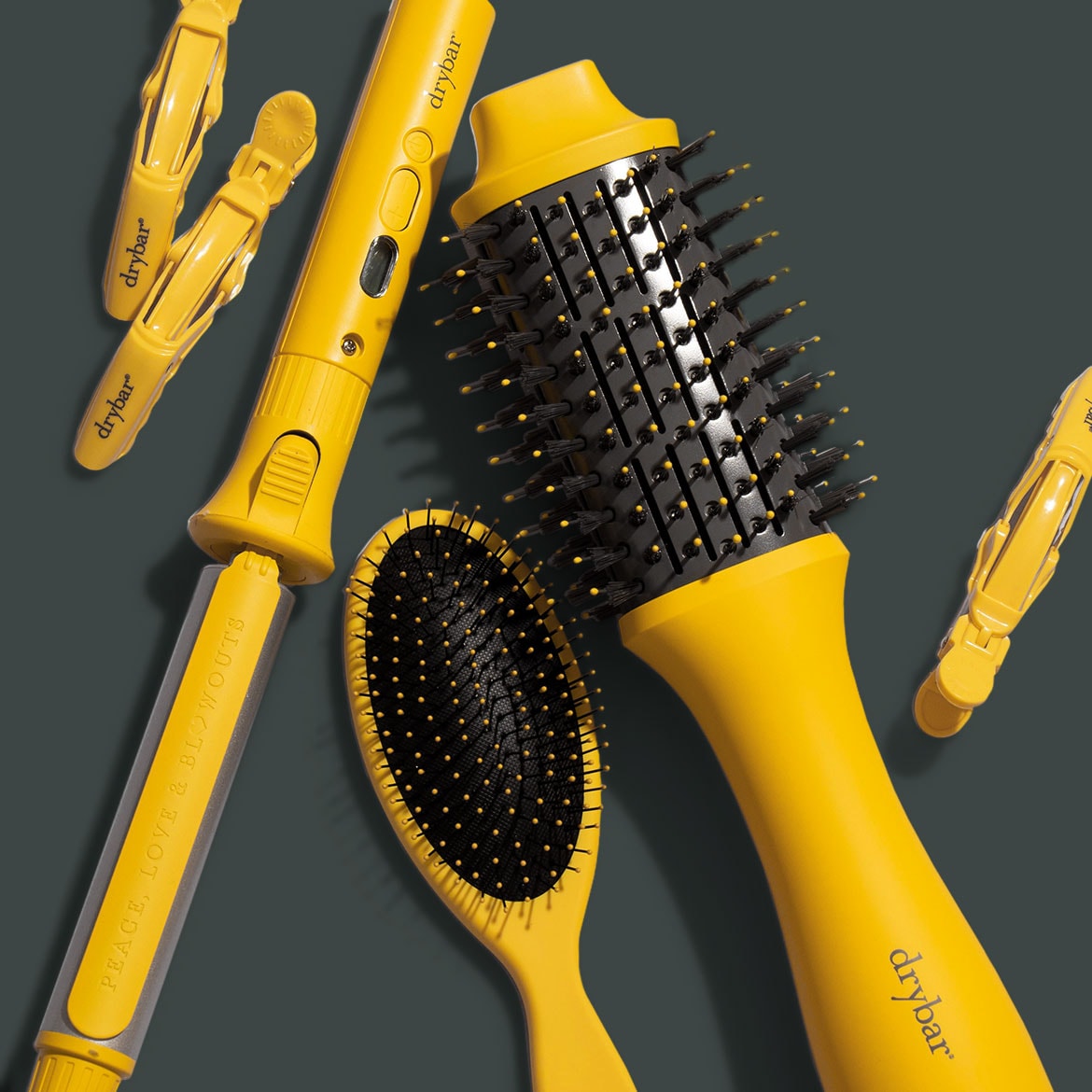 Gele Drybar-stylingtools, waaronder een krultang, borstels en clips, liggen op een grijze achtergrond.