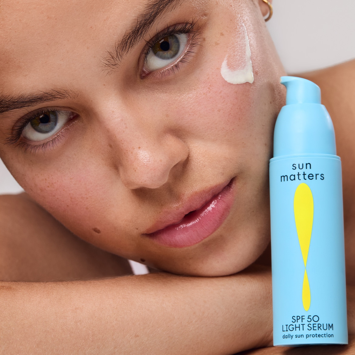 Een vrouw met een druppel Sun Matters SPF 50 Light Serum op haar gezicht, de lichtblauwe verpakking belooft dagelijkse bescherming.