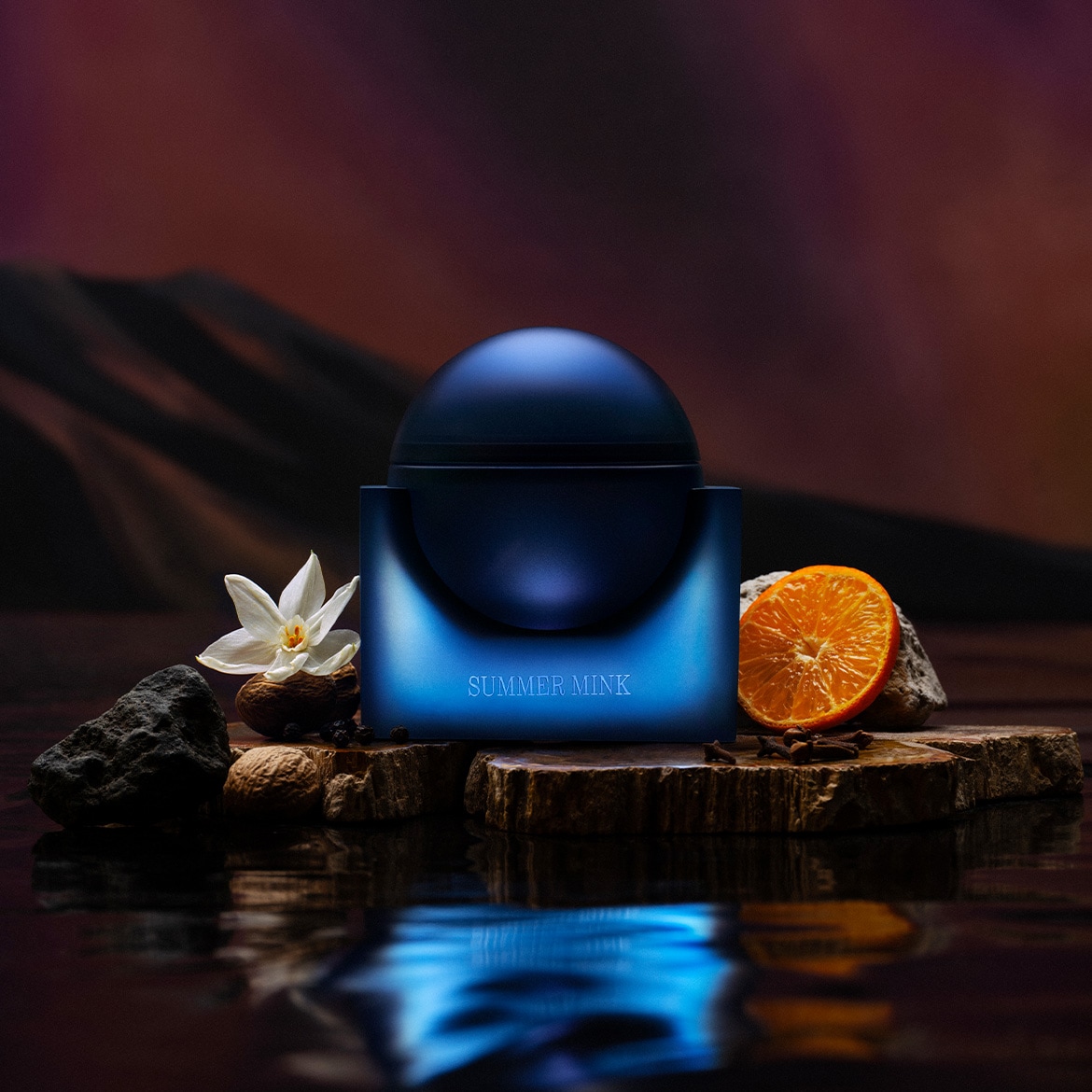 Een donkerblauwe, bolvormige pot met de tekst "SUMMER MINK" staat op een houten plank, omringd door een witte bloem, noten, rotsen en een sinaasappelschijfje, met een reflectie in het water eronder.