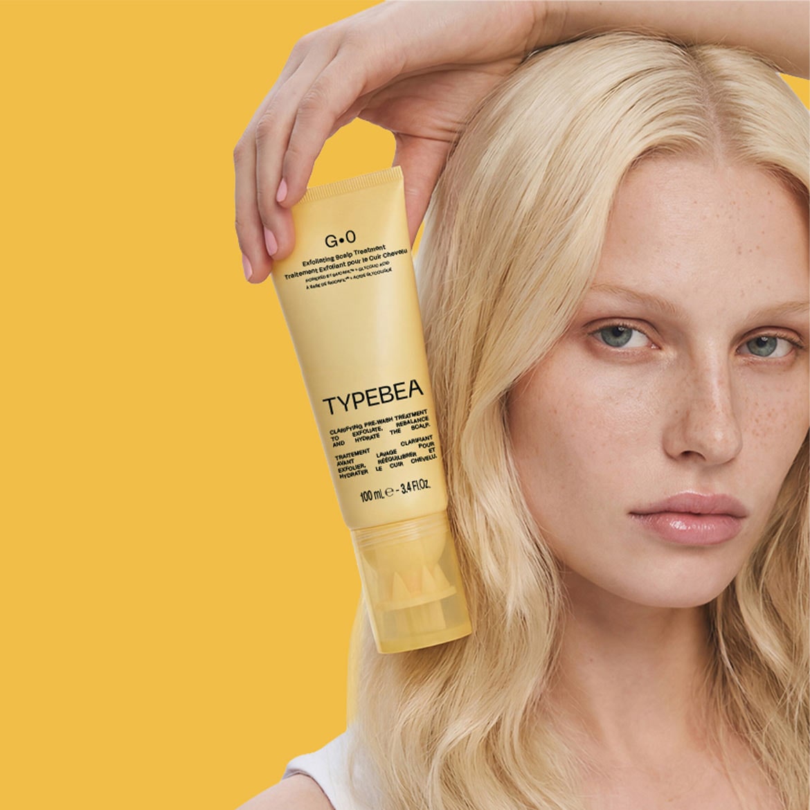 Een vrouw met blond haar houdt een gele tube G.O Exfoliating Scalp Treatment van Typebea vast, een zuiverende pre-wash behandeling van 100 ml die de hoofdhuid exfolieert, hydrateert en in balans brengt.