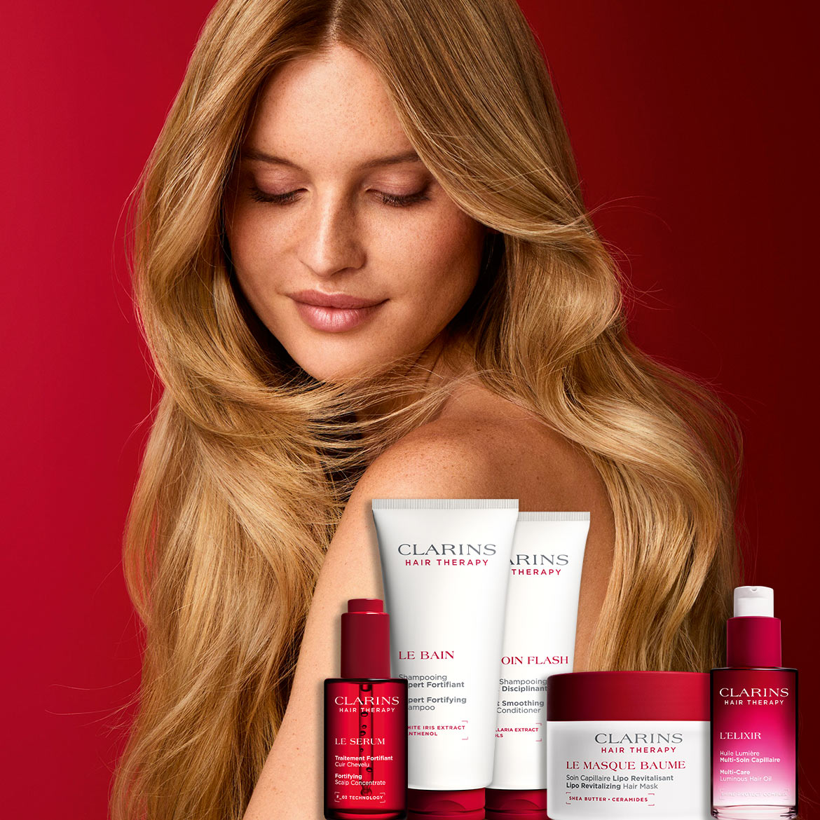 Een vrouw met lang, blond haar en een natuurlijke make-up poseert met een assortiment Clarins Hair Therapy producten, waaronder een shampoo, conditioner, serum, masker en haarolie, allemaal in rood-witte verpakkingen.