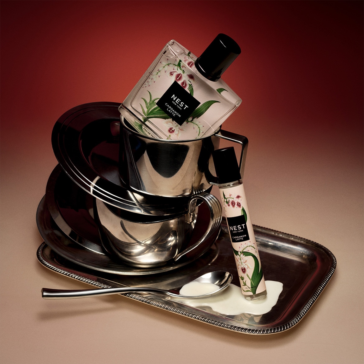 De Nest New York Cardamom Latte Eau de Parfum, een vierkante fles met bloemmotief, staat op een stapel zilveren kopjes en schoteltjes, met een kleinere rollerfles ernaast op een zilveren dienblad met gemorste witte vloeistof en een lepel.