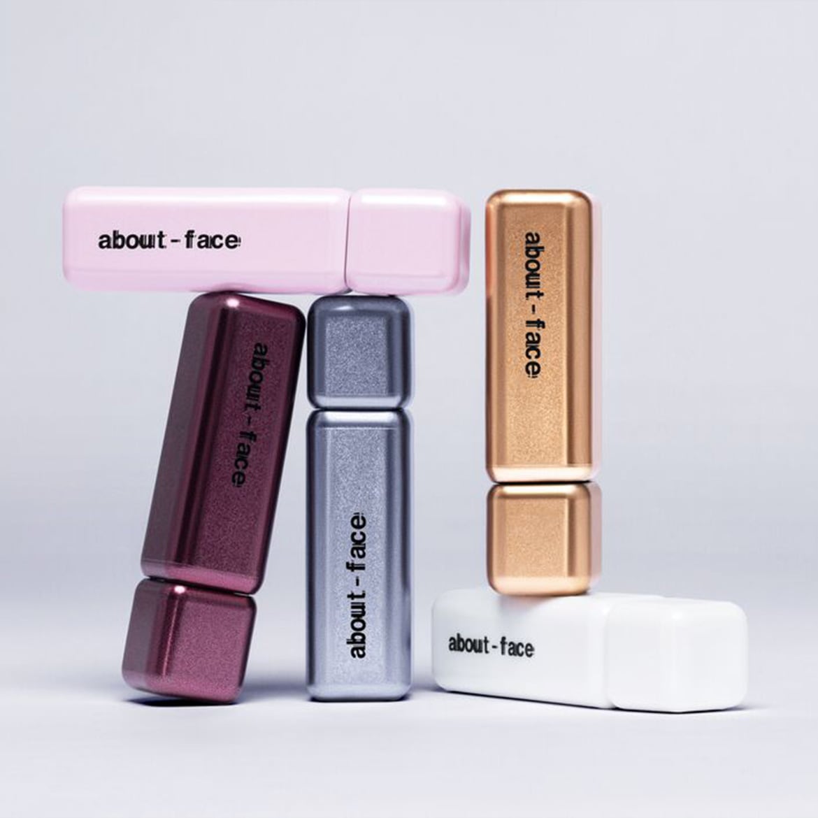 Vijf About-Face make-up producten in verschillende kleuren, waaronder roze, bordeauxrood, zilver, goud en wit, zijn artistiek gestapeld op een lichte achtergrond.