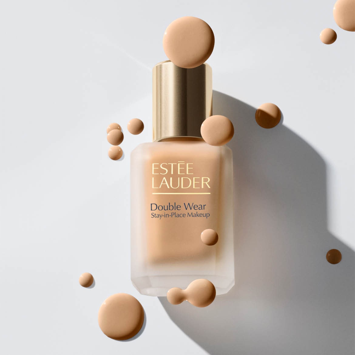 Ontdek Estée Lauder Double Wear Stay-in-Place Makeup, een matte foundation in een glazen fles met gouden dop, omringd door druppels foundation.