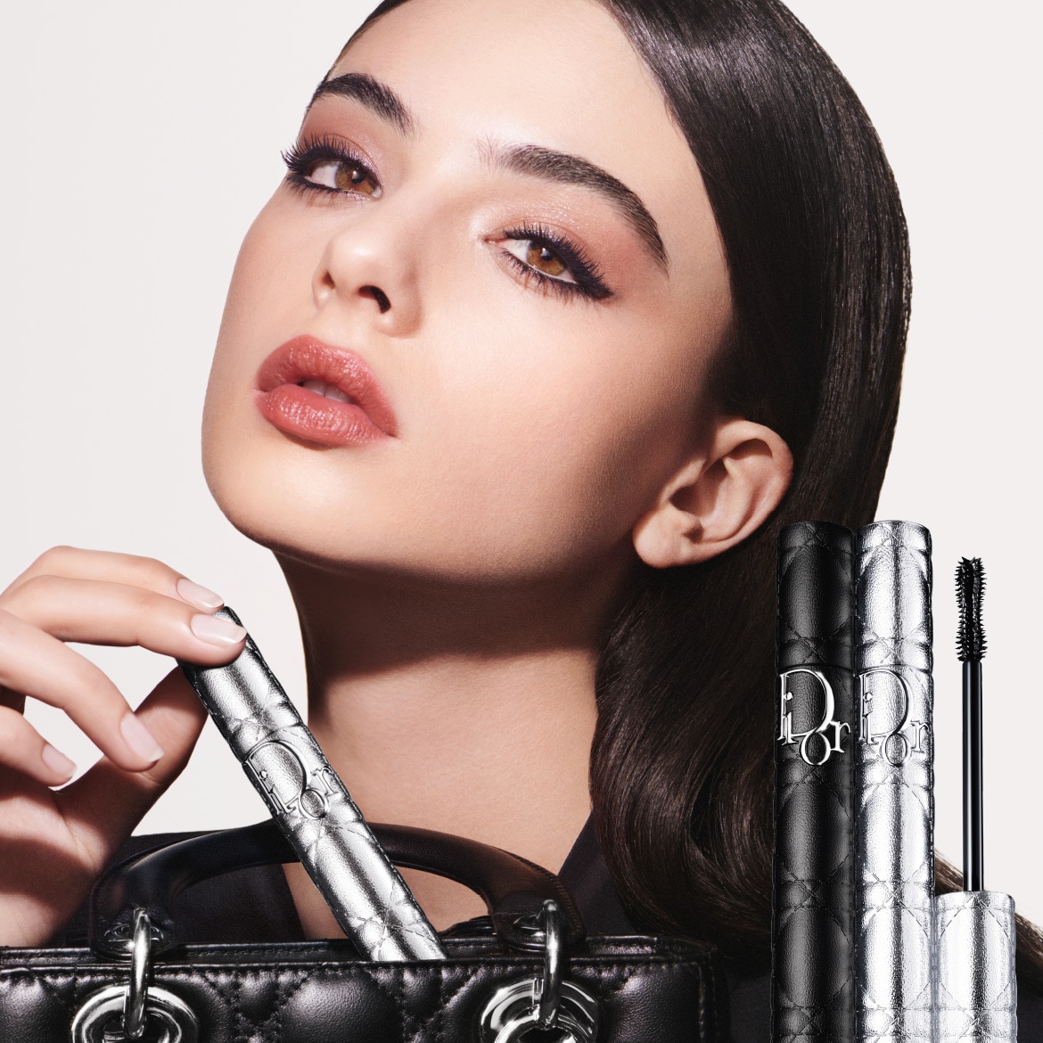 Close-up van een vrouw met bruine ogen en een perzikkleurige lippenstift, die een zilveren Dior mascara vasthoudt, met de mascara verpakking op de voorgrond.