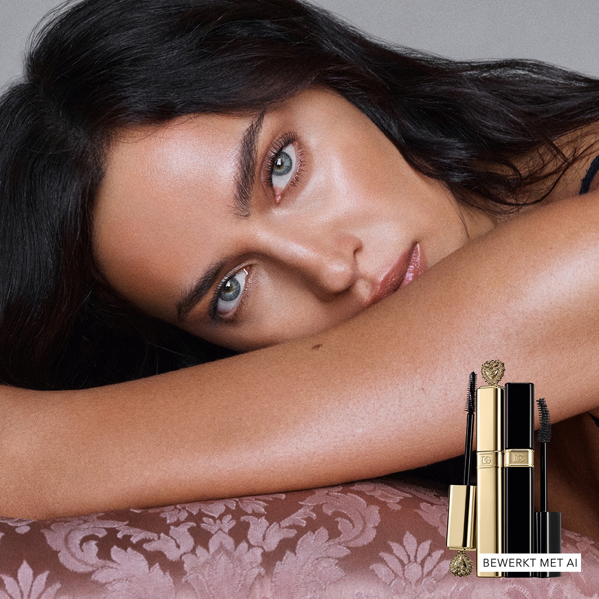 Een vrouw met donker haar en helderblauwe ogen leunt op haar arm, met op de voorgrond twee Dolce & Gabbana mascara's in goud en zwart, met de tekst "BEWERKT MET AI" in de hoek.