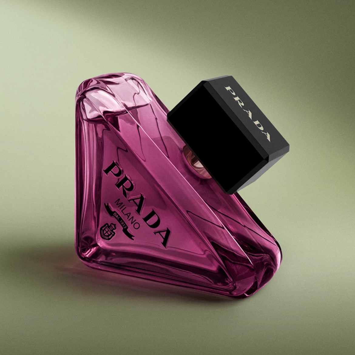 De Prada Paradoxe Intense Eau de Parfum, een roze, driehoekige flacon met een zwarte dop, ligt schuin op een lichtgroene achtergrond.