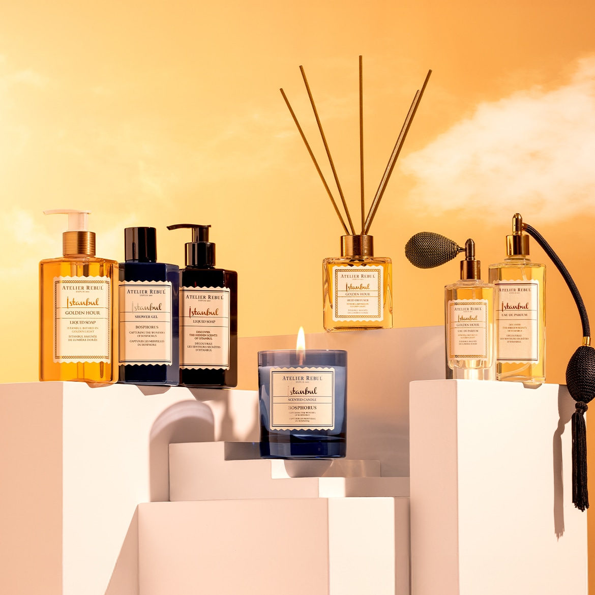 Een collectie Atelier Rebul Istanbul producten, waaronder vloeibare zeep, douchegel, een geurdiffuser, parfum en een geurkaars, gepresenteerd op witte platforms tegen een oranje lucht.