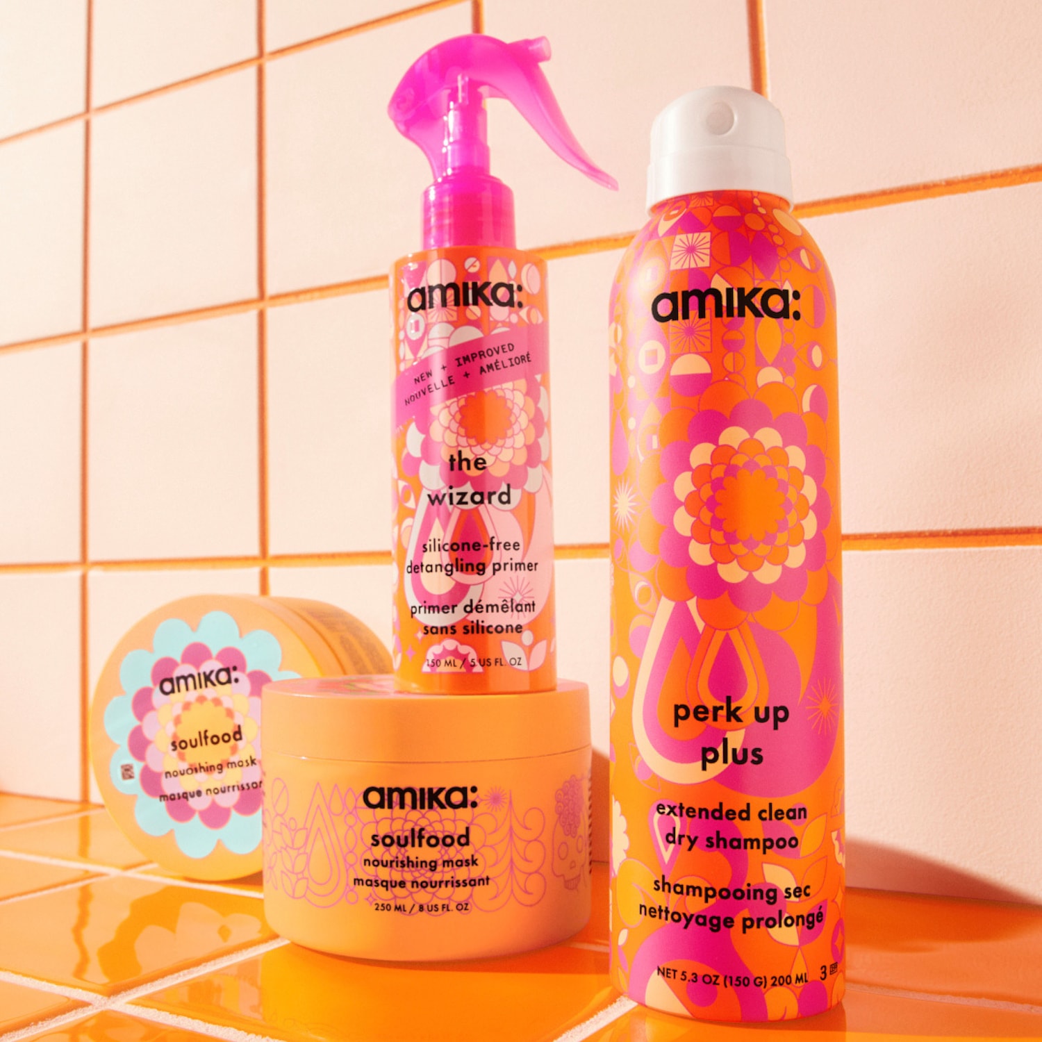 Vier Amika haarverzorgingsproducten staan op een oranje tegelachtergrond, waaronder The Wizard ontwarrende primer, Soulfood voedend masker en Perk Up Plus droogshampoo, allemaal in felgekleurde verpakkingen.