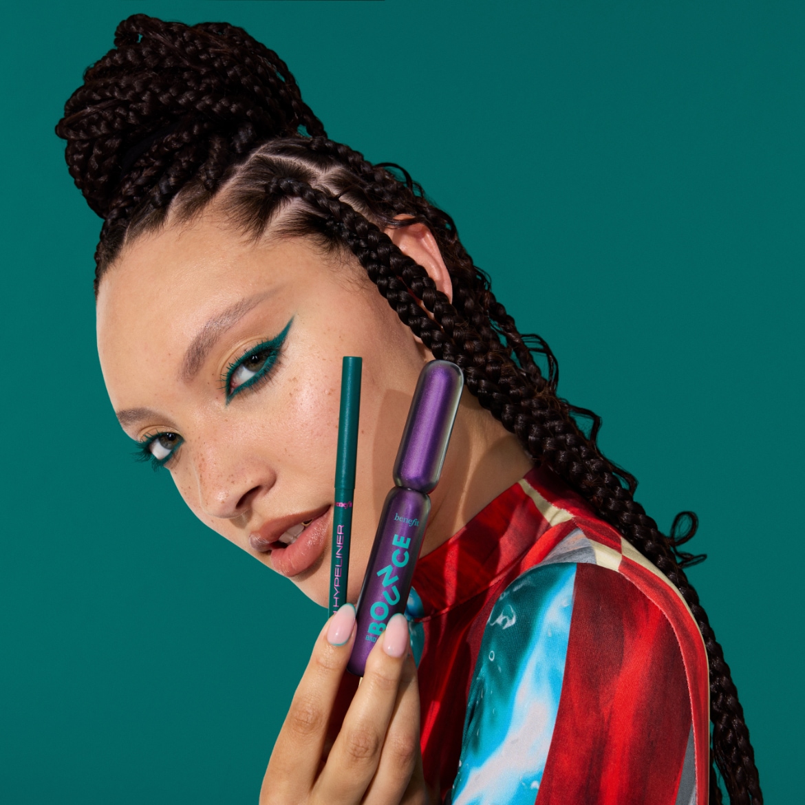 Een vrouw met gevlochten haar en een kleurrijke top houdt een groene eyeliner en een paarse mascara vast, met haar ogen opgemaakt met een bijpassende groene eyeliner.