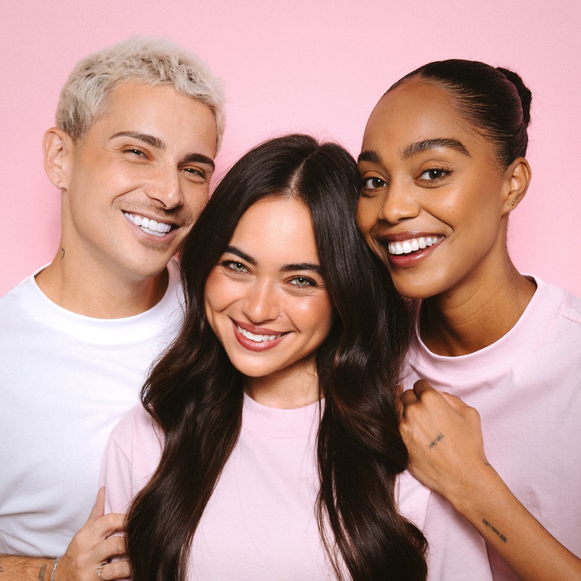 Drie lachende modellen, twee vrouwen en één man, met een stralende huid en tanden, poseren tegen een roze achtergrond, waarbij de man en een vrouw witte T-shirts dragen en de andere vrouw een roze T-shirt.