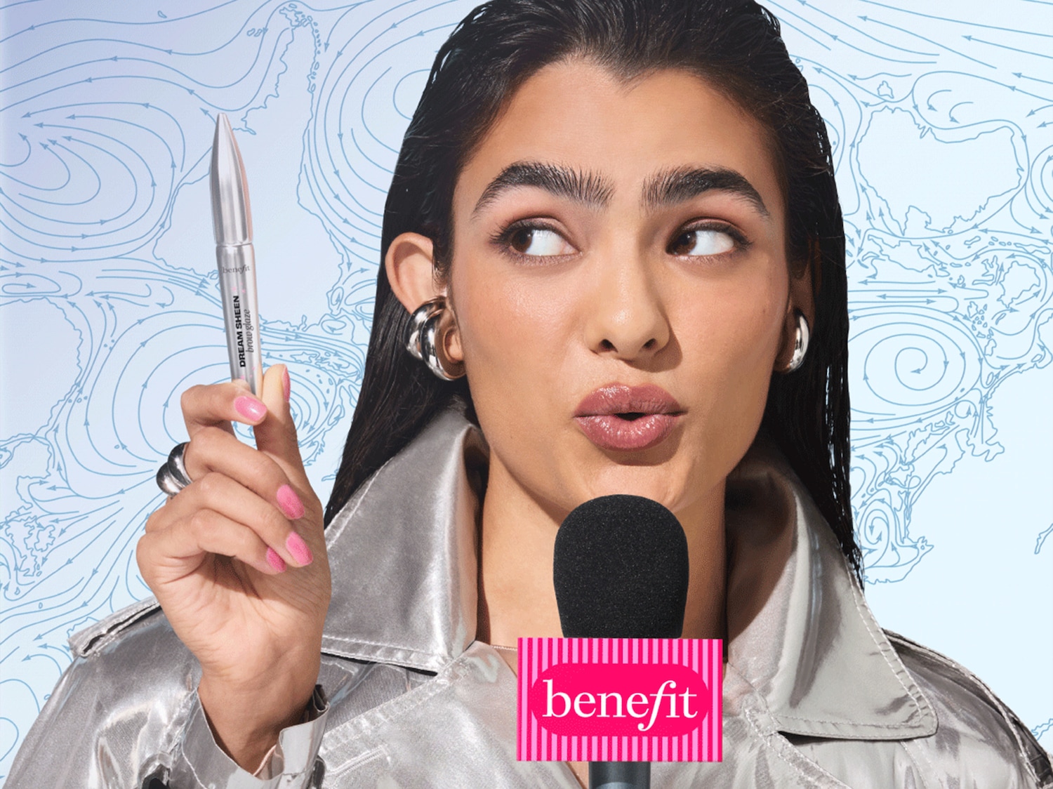 Een vrouw met een zilverkleurige jas en roze nagellak houdt een Benefit Dream Sheen Brow Glaze vast, terwijl ze in een microfoon met het Benefit-logo spreekt, tegen een achtergrond van een weerkaart.