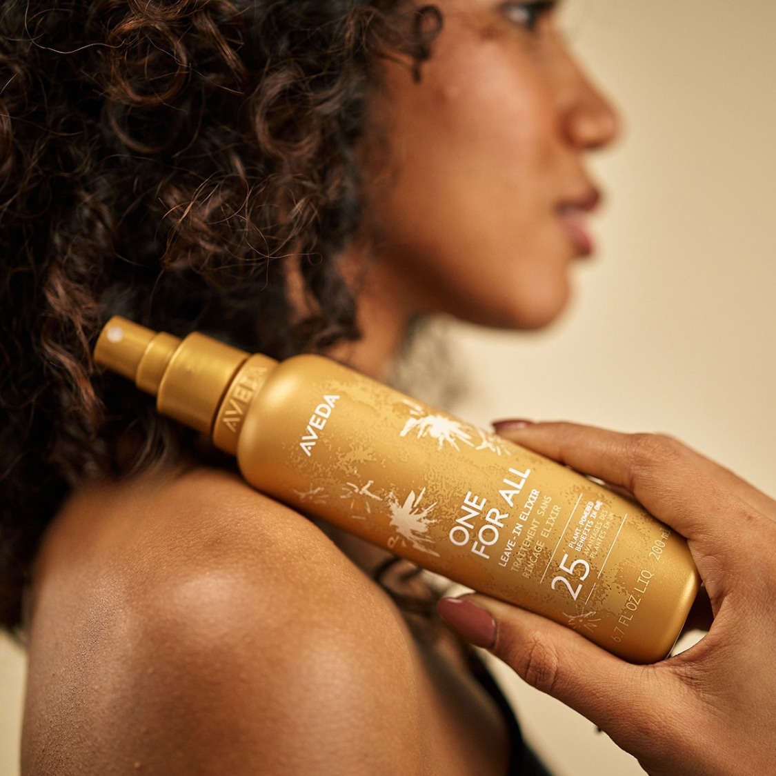 Een vrouw met krullend haar houdt een goudkleurige fles Aveda One For All Leave-In Elixir vast, met de tekst "25 plant-powered benefits in one" erop.