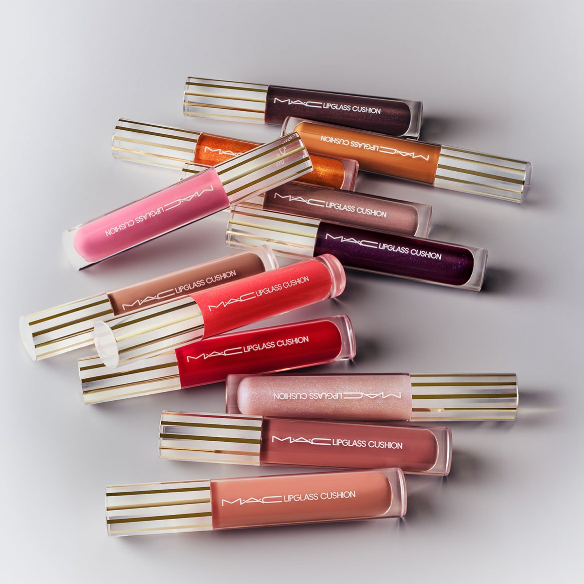 Een verzameling MAC Lipglass Cushion lipglosses in verschillende kleuren, van roze en rood tot bruin en paars, allemaal met een transparante verpakking met gouden strepen.