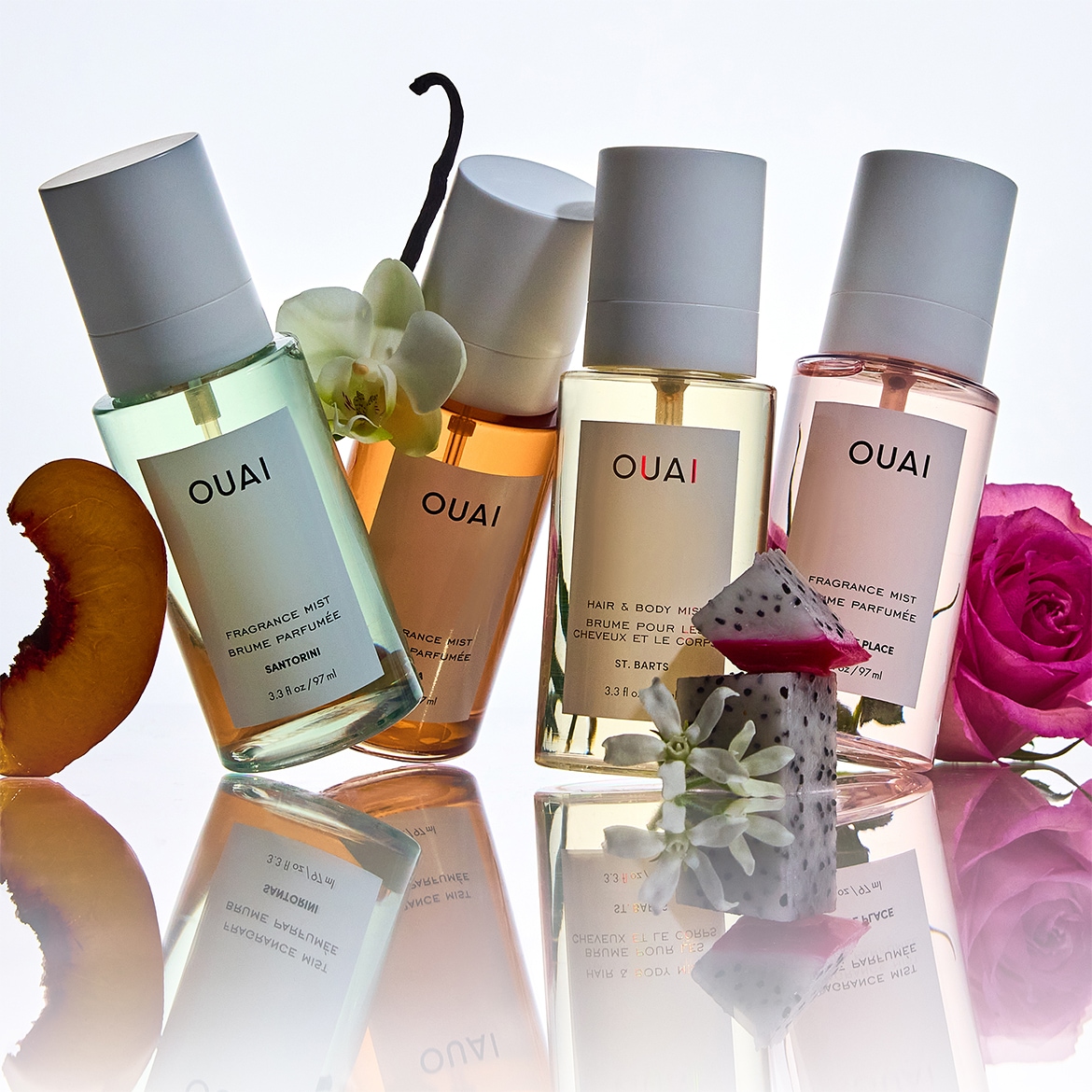 Vier flessen OUAI-parfumnevel in verschillende kleuren, elk met een unieke geur, gecombineerd met fruit en bloemen die de geuren vertegenwoordigen.