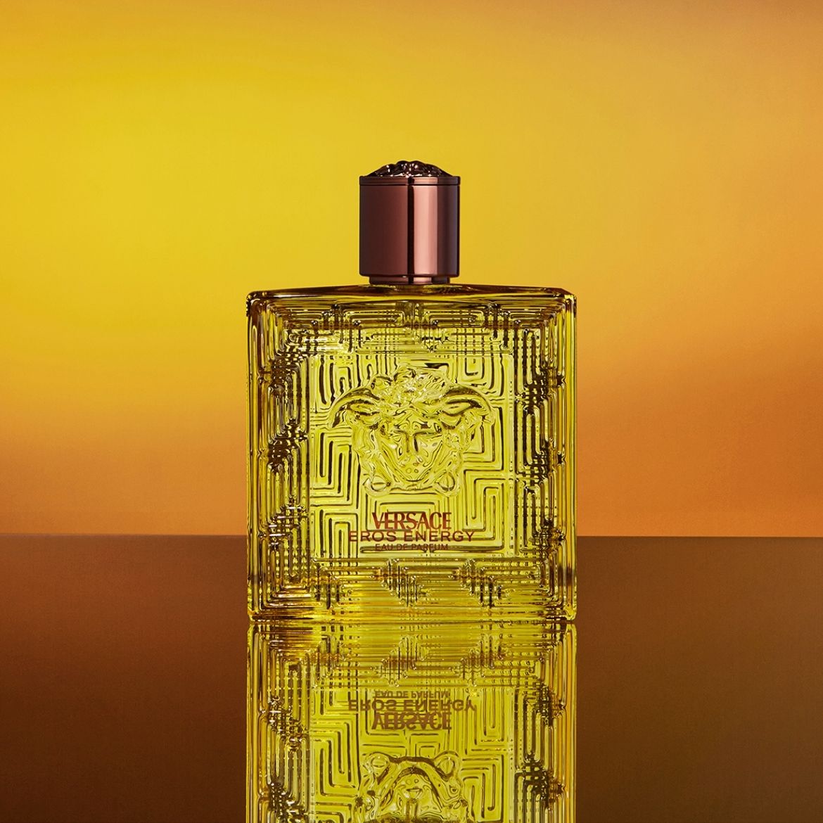 Gouden vierkante Versace Eros Energy Eau de Parfum fles met een bruine dop, versierd met een Grieks sleutelmotief en een Medusa-logo, tegen een gele achtergrond.