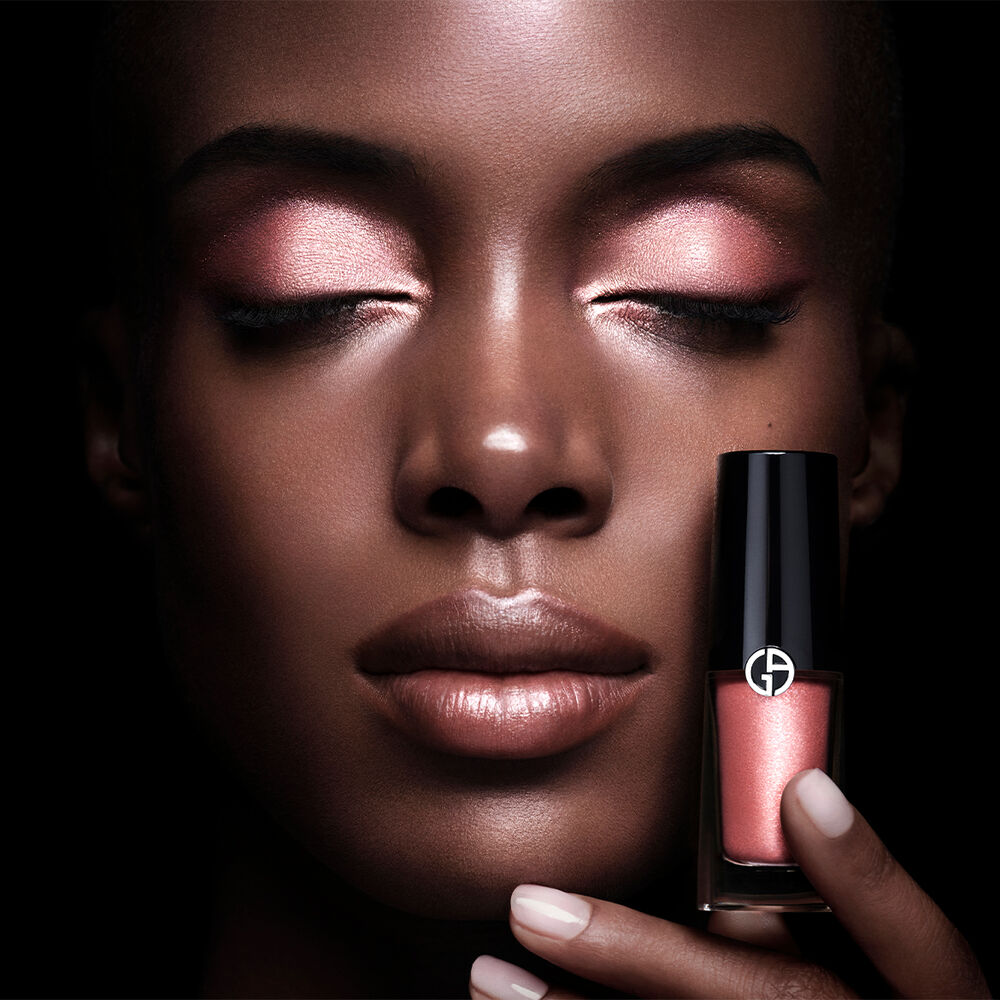 Een vrouw met een donkere huidskleur en glanzende, perzikkleurige oogschaduw en lippen, houdt een flesje nagellak in een bijpassende perzikkleurige tint vast.