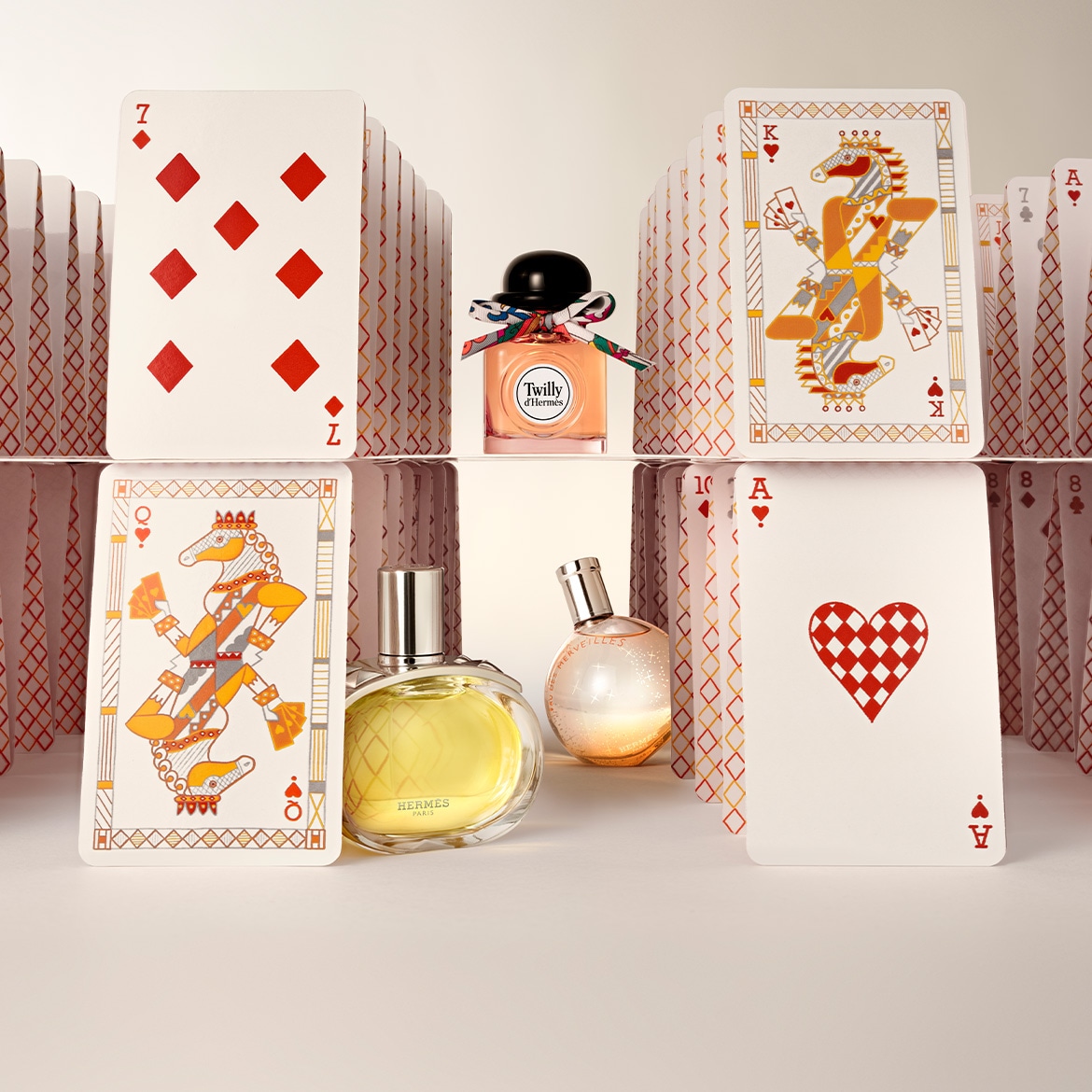 De foto toont een collectie Hermès-parfums, waaronder Twilly d'Hermès met een kleurrijk lint, geplaatst tussen speelkaarten zoals zevens, heren en azen.