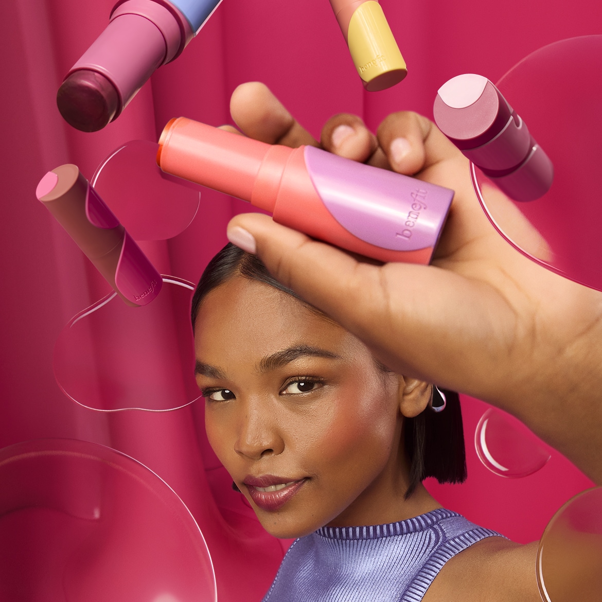 Een vrouw houdt een Benefit blush stick vast, omringd door andere blush sticks in roze, perzik en bruine tinten, tegen een roze achtergrond.