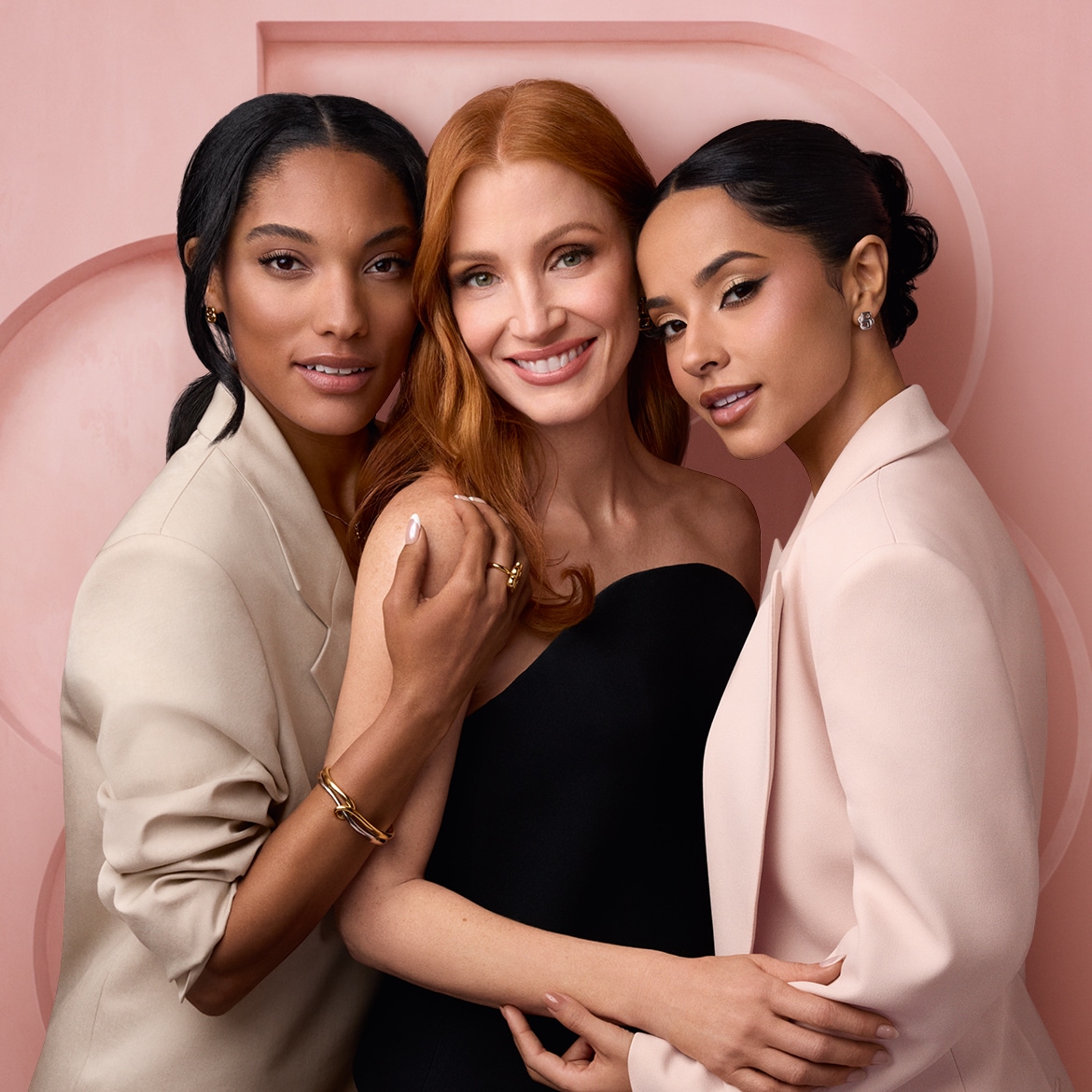 Drie vrouwen, waaronder Jessica Chastain, poseren lachend voor een roze achtergrond, waarbij de middelste vrouw een zwarte strapless jurk draagt en de andere twee blazers in beige en lichtroze.