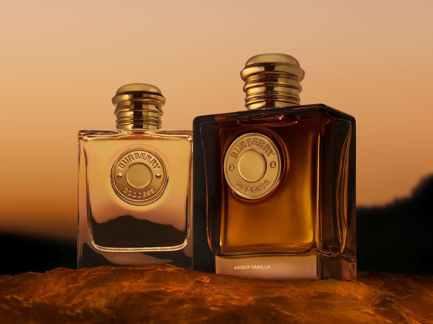 Twee vierkante Burberry Goddess parfumflessen met gouden doppen en labels, de linker fles is licht amberkleurig en de rechter fles is donker amberkleurig, staan op een golvend, oranjebruin oppervlak tegen een oranje zonsondergang.