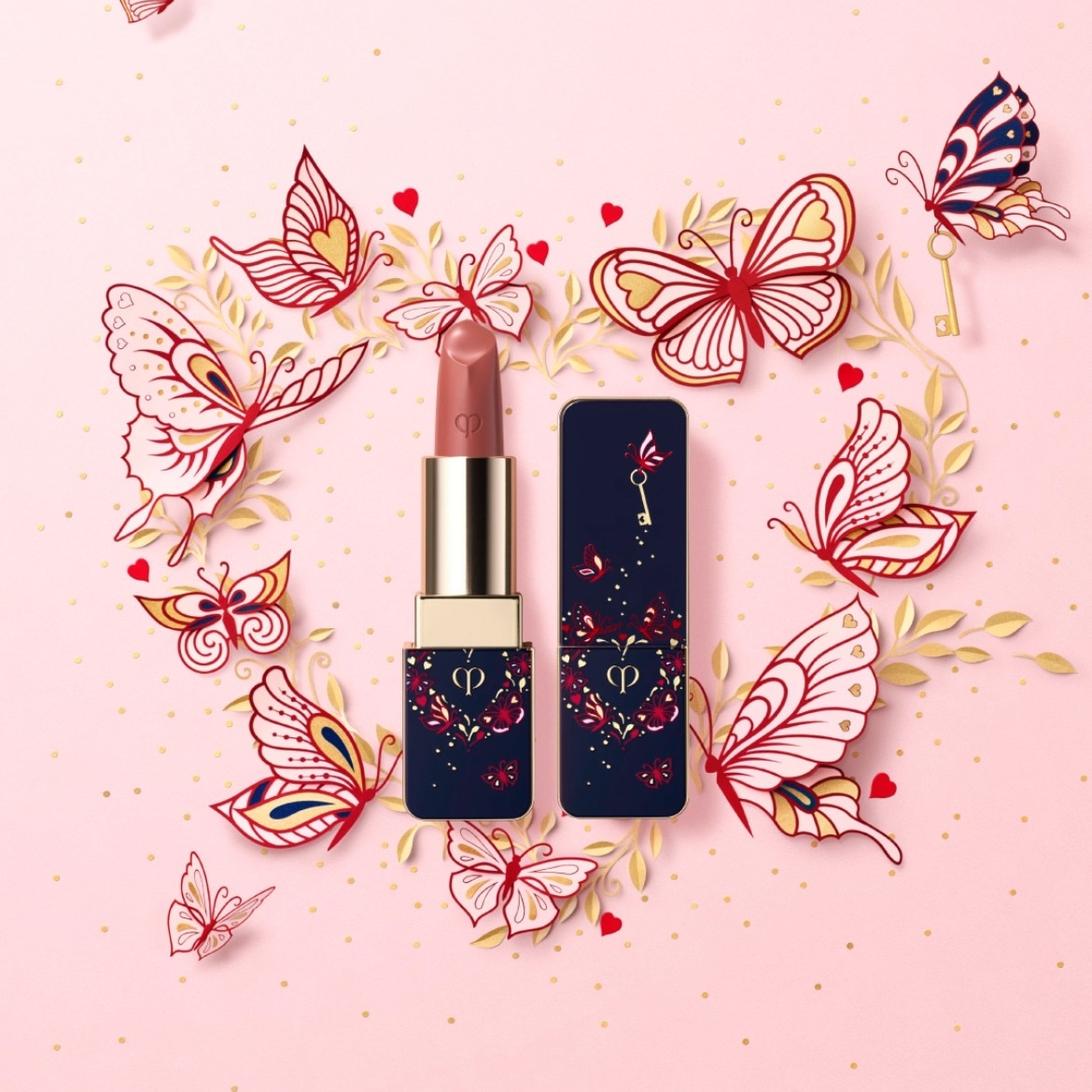Een nude kleurige lippenstift van Clé de Peau, omringd door papieren vlinders en gouden stippen, staat naast de donkerblauwe verpakking met een gouden sleutel.