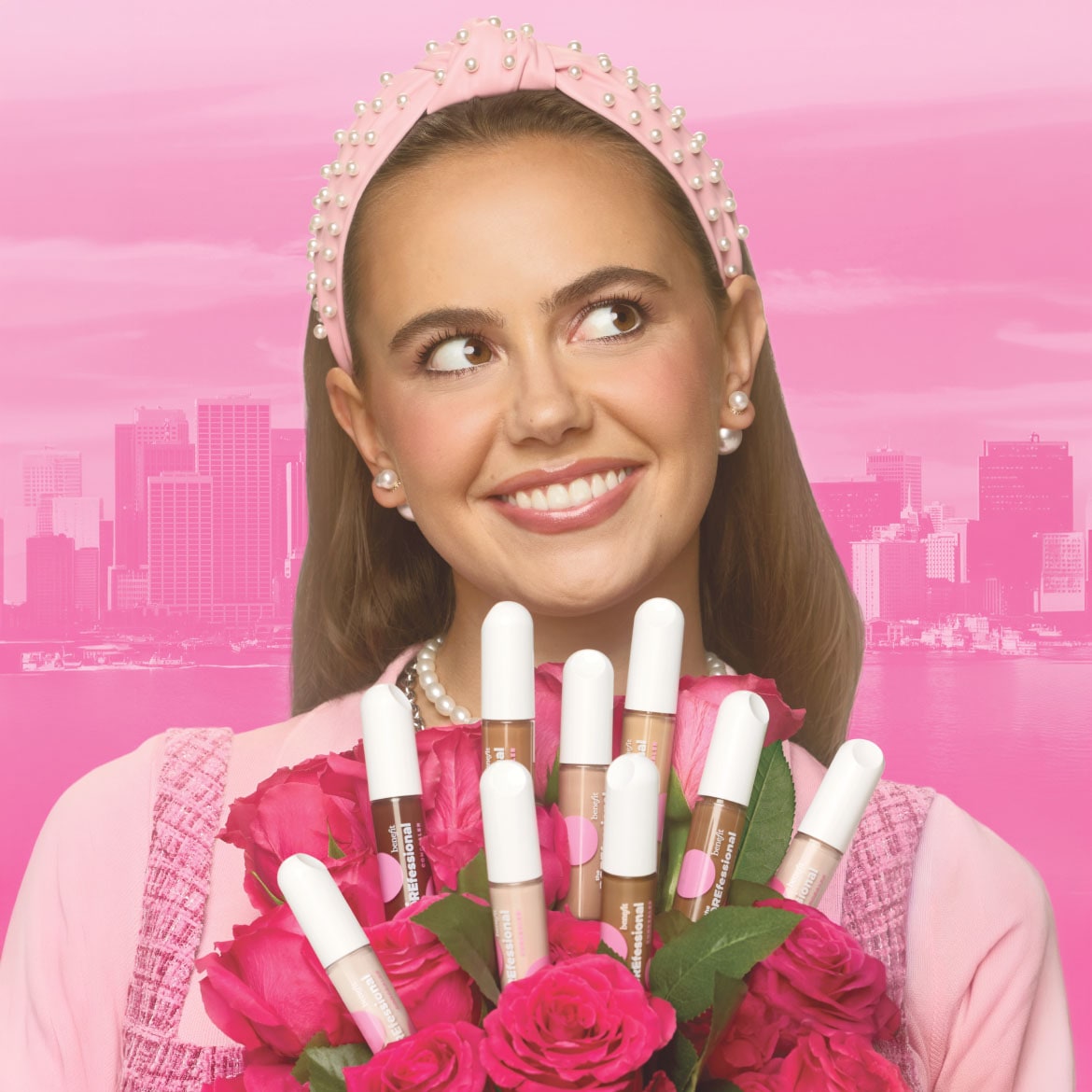 Een lachende vrouw met een roze haarband met parels en een parelketting, houdt een boeket roze rozen vast met daarin verschillende Benefit Professional concealers in diverse huidtinten, tegen een roze achtergrond met een stadsskyline.