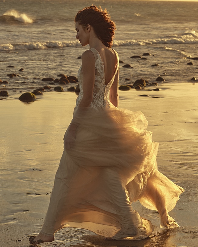 Een vrouw in een lange, vloeiende jurk met kanten details loopt langs het strand bij zonsondergang, de golven zachtjes op de achtergrond.