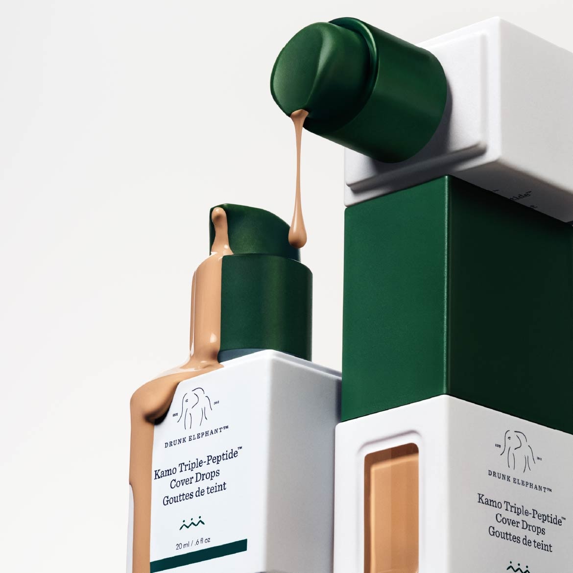 Drunk Elephant Kamo Triple-Peptide Cover Drops in een witte fles met donkergroene dop, waaruit een beige vloeistof druppelt, staat op een stapel verpakkingen.