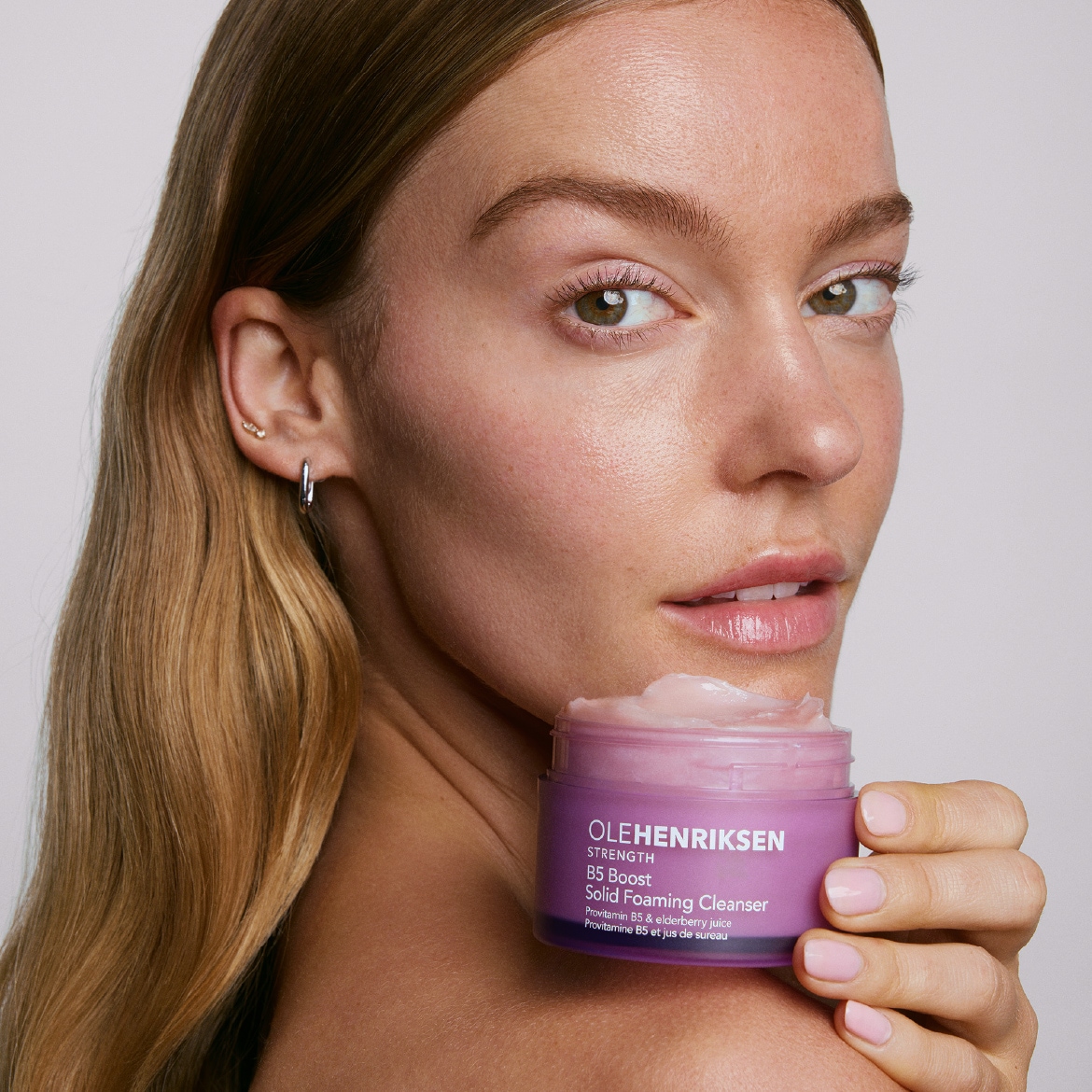 Een vrouw met lang blond haar en een natuurlijke make-uplook houdt een paarse pot OLEHENRIKSEN Strength B5 Boost Solid Foaming Cleanser vast, met een lichtroze, romige textuur zichtbaar.