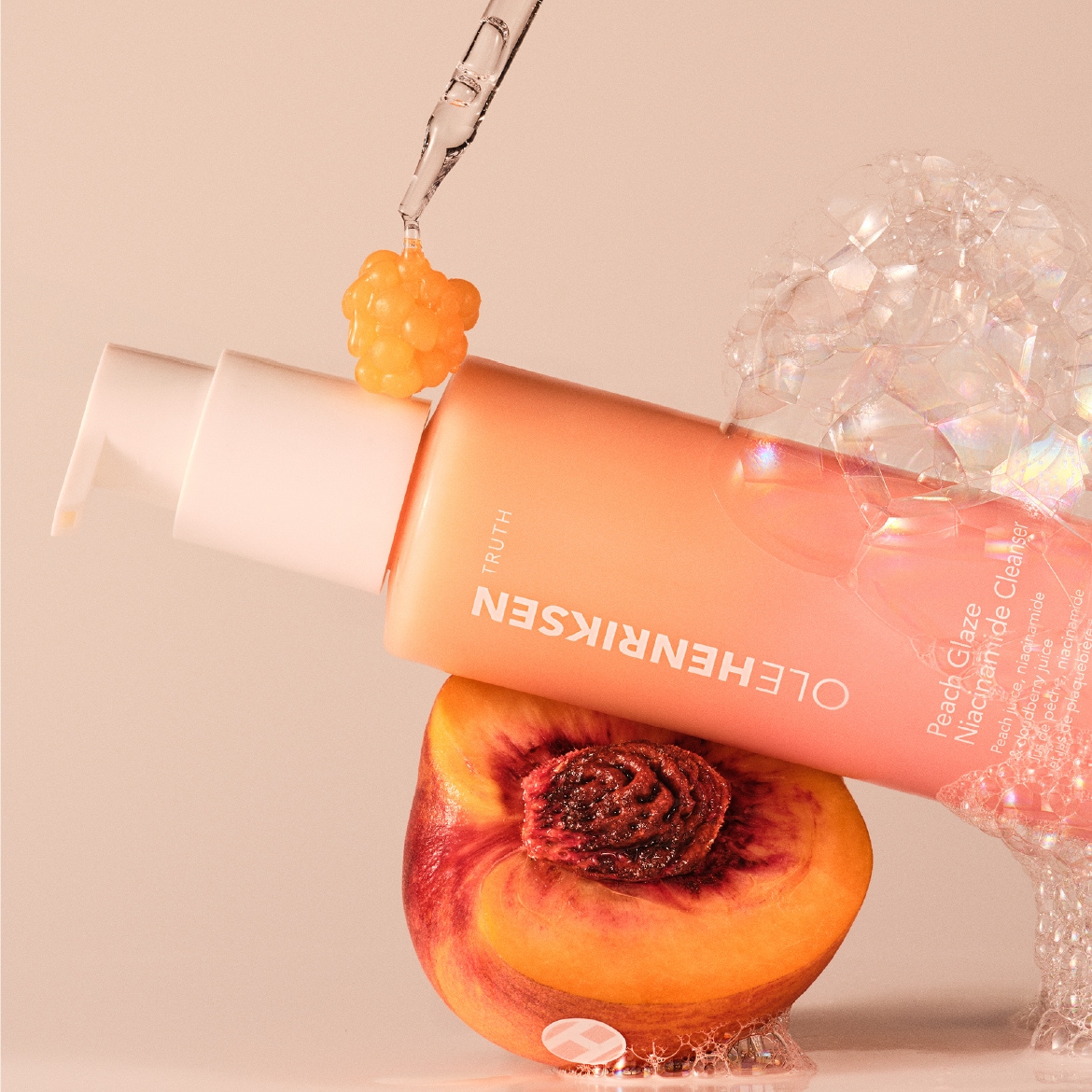 De Ole Henriksen Peach Glaze Niacinamide Cleanser in een perzikkleurige verpakking ligt op een halve perzik, met een druppel serum erboven en bubbels eromheen.
