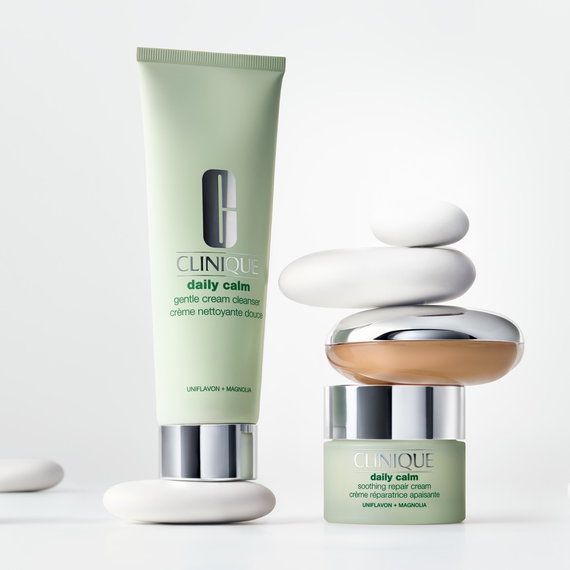 De Clinique Daily Calm Gentle Cream Cleanser en Soothing Repair Cream, beide in lichtgroene verpakkingen, staan op gestapelde witte stenen, met een extra bruine pot ertussen.