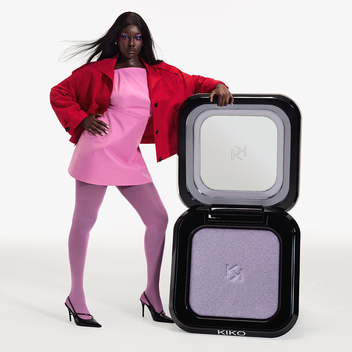 Een model met donkere huidskleur, gekleed in een roze jurk, rode jas en paarse panty, poseert naast een open, vierkante zwarte make-updoos met een spiegel en lila oogschaduw.