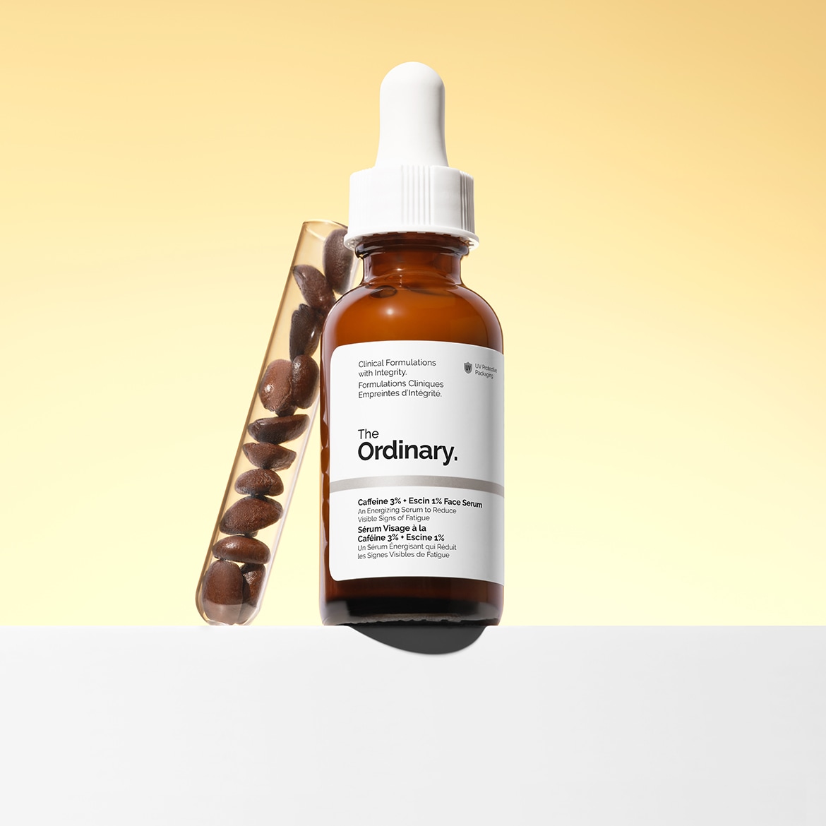 De Ordinary Caffeine 3% + Escin 1% Face Serum in een bruine glazen fles met een witte pipetdop, gepresenteerd naast een reageerbuis gevuld met koffiebonen.