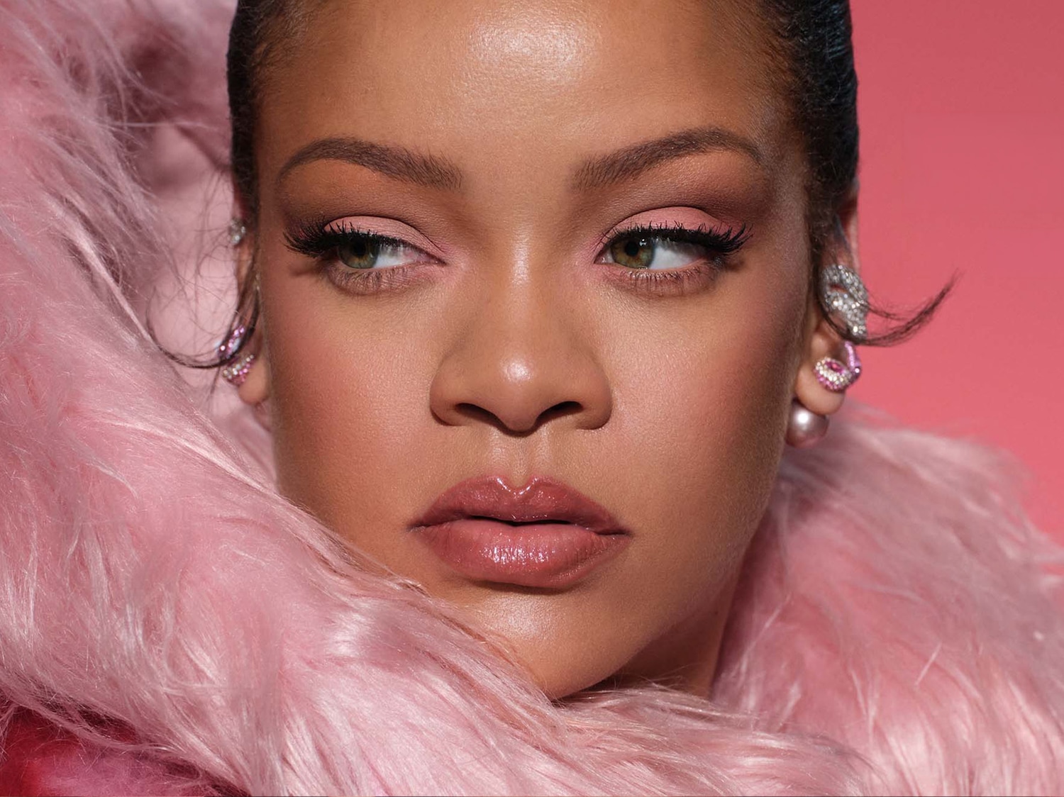 Rihanna draagt een roze, pluizige jas en heeft een natuurlijke make-uplook met roze oogschaduw en glanzende lippen.