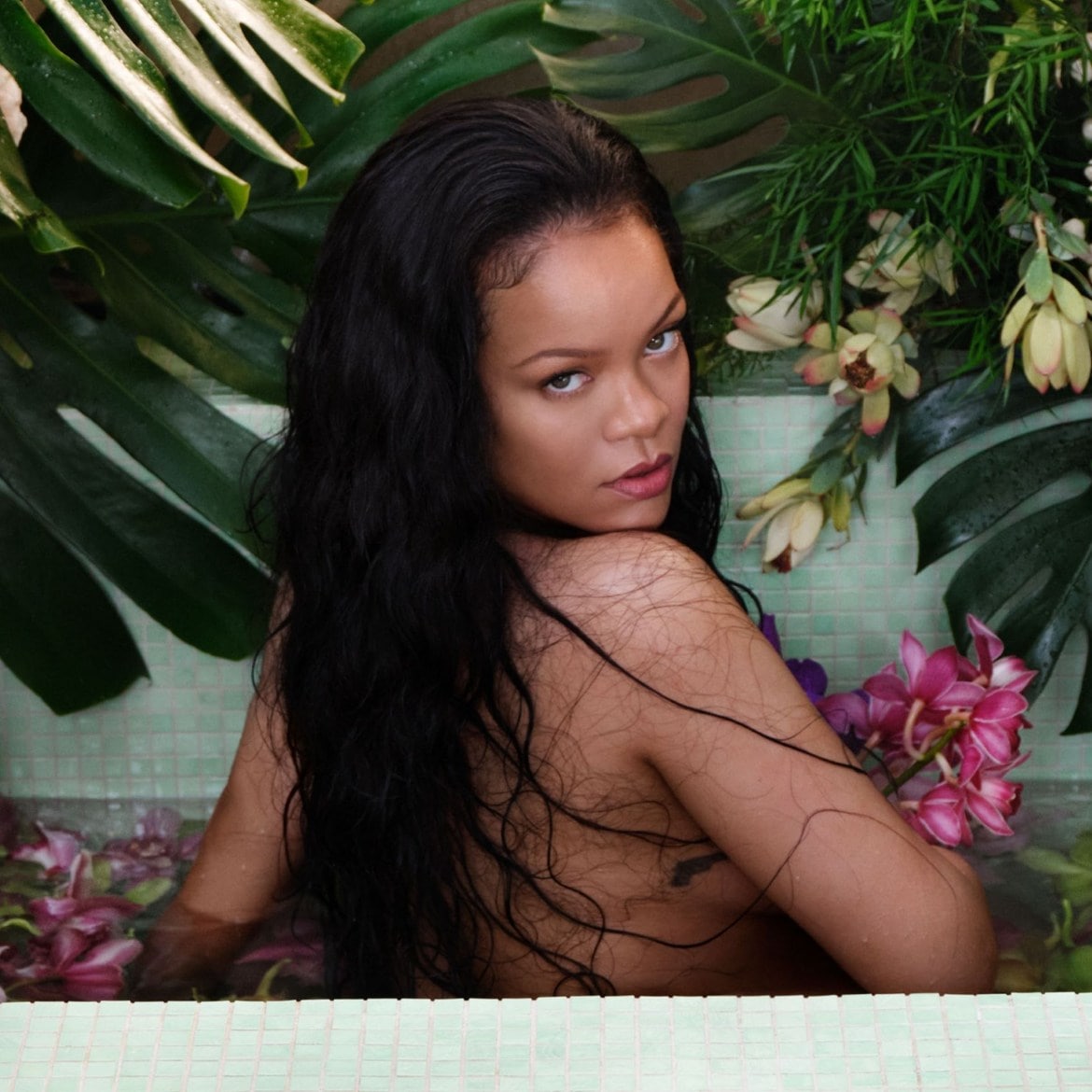 Rihanna poseert in een bad vol water en bloemen, met weelderig groen op de achtergrond, haar donkere haar valt over haar schouders.