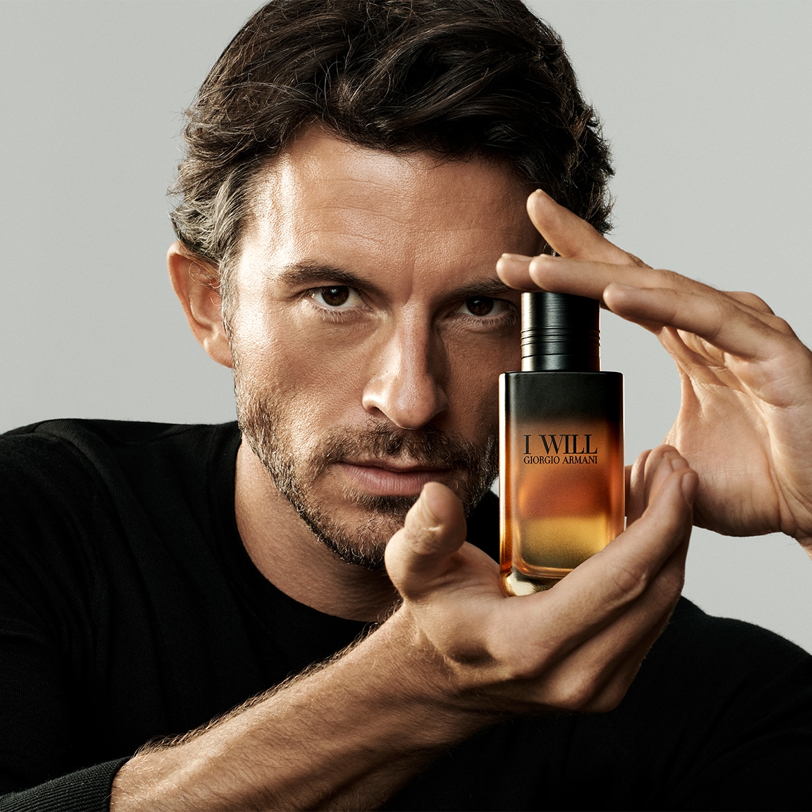 Een man met donker haar en een lichte baard houdt een Giorgio Armani "I WILL" parfumflesje vast, met een donkere dop en een oranje-gele vloeistof, tegen een grijze achtergrond.