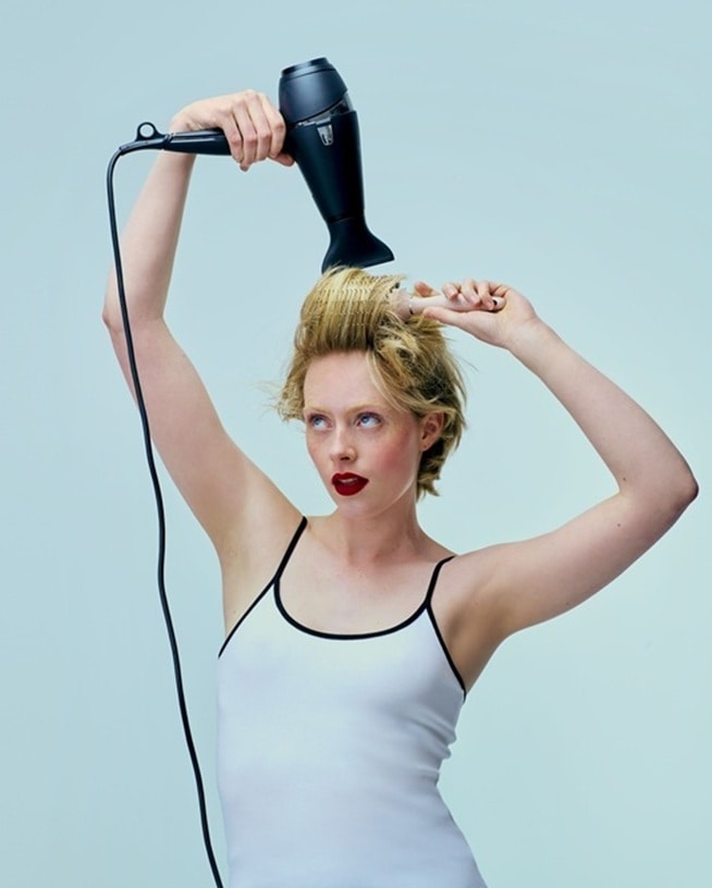 Een vrouw met rode lippenstift gebruikt een zwarte föhn en een borstel om haar blonde haar te stylen, waarmee ze een perfecte styling creëert zonder schade door de hitte.'