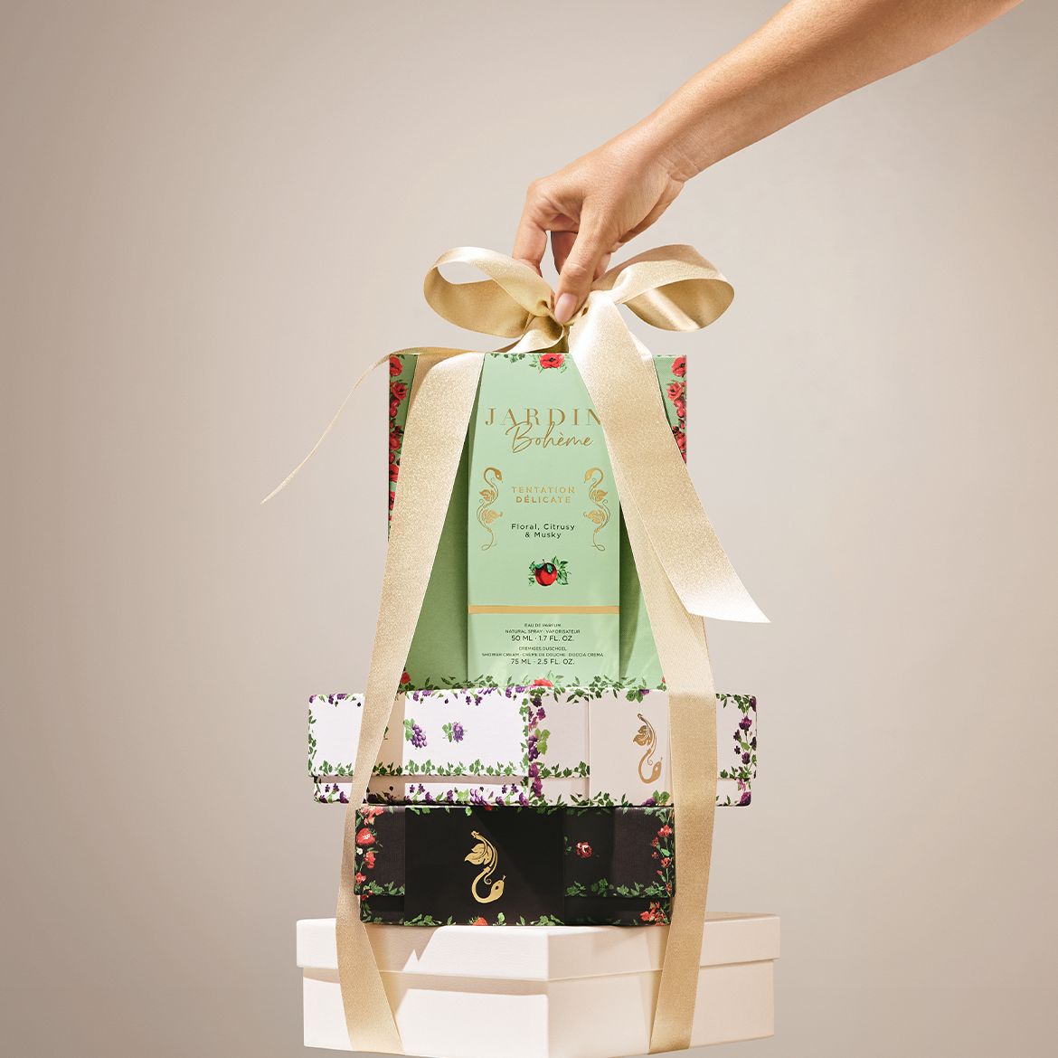 Een hand houdt een geschenkset van Jardin Bohème Tentation Délicate vast, met een lichtgroene doos met bloemen, citrus en muskusgeuren, en daaronder twee kleinere dozen met bloemmotieven, allemaal samengebonden met een gouden lint.