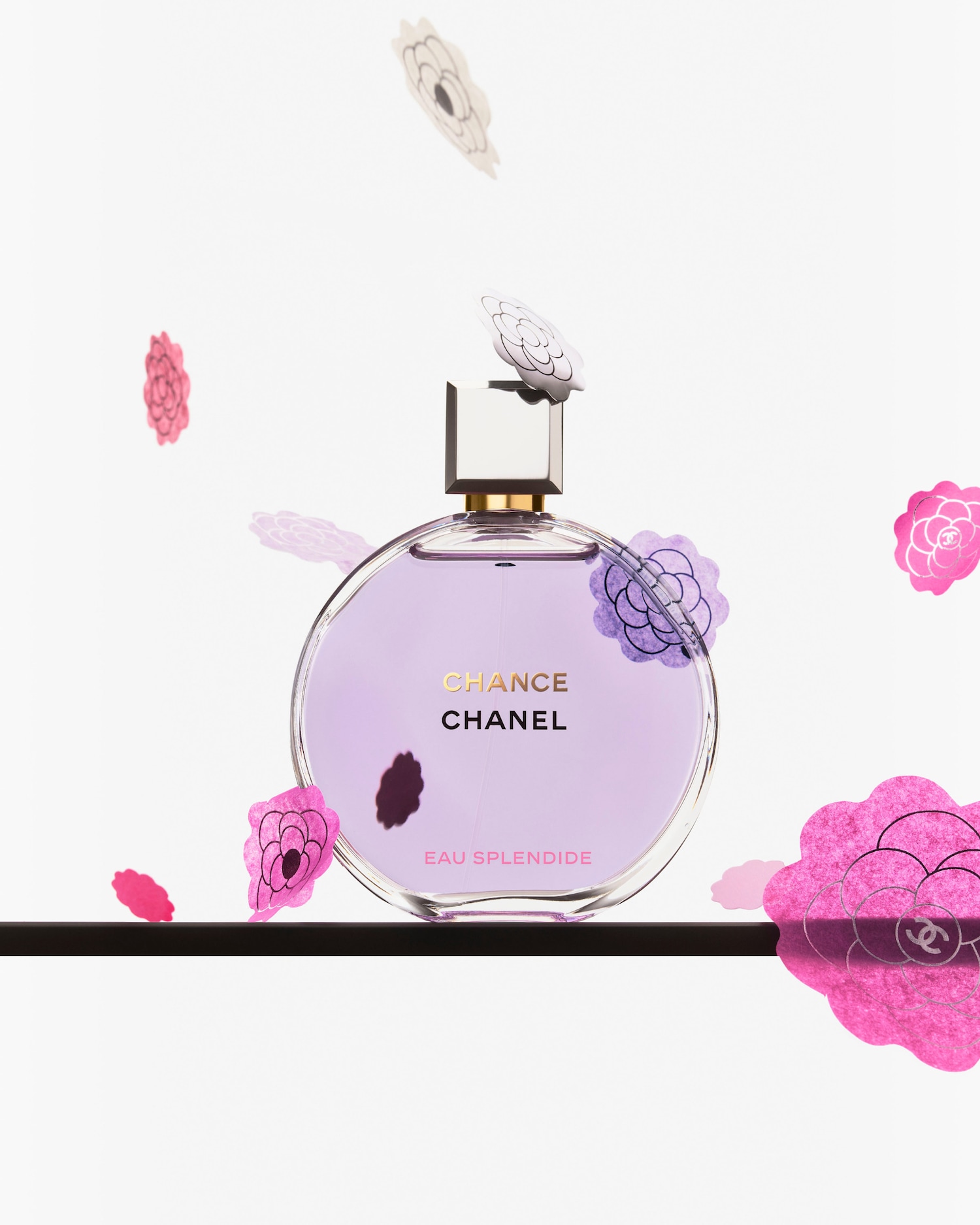 Een ronde, lichtpaarse Chanel Chance Eau Splendide parfumfles staat op een zwarte plank, omringd door roze en paarse bloemdecoraties.