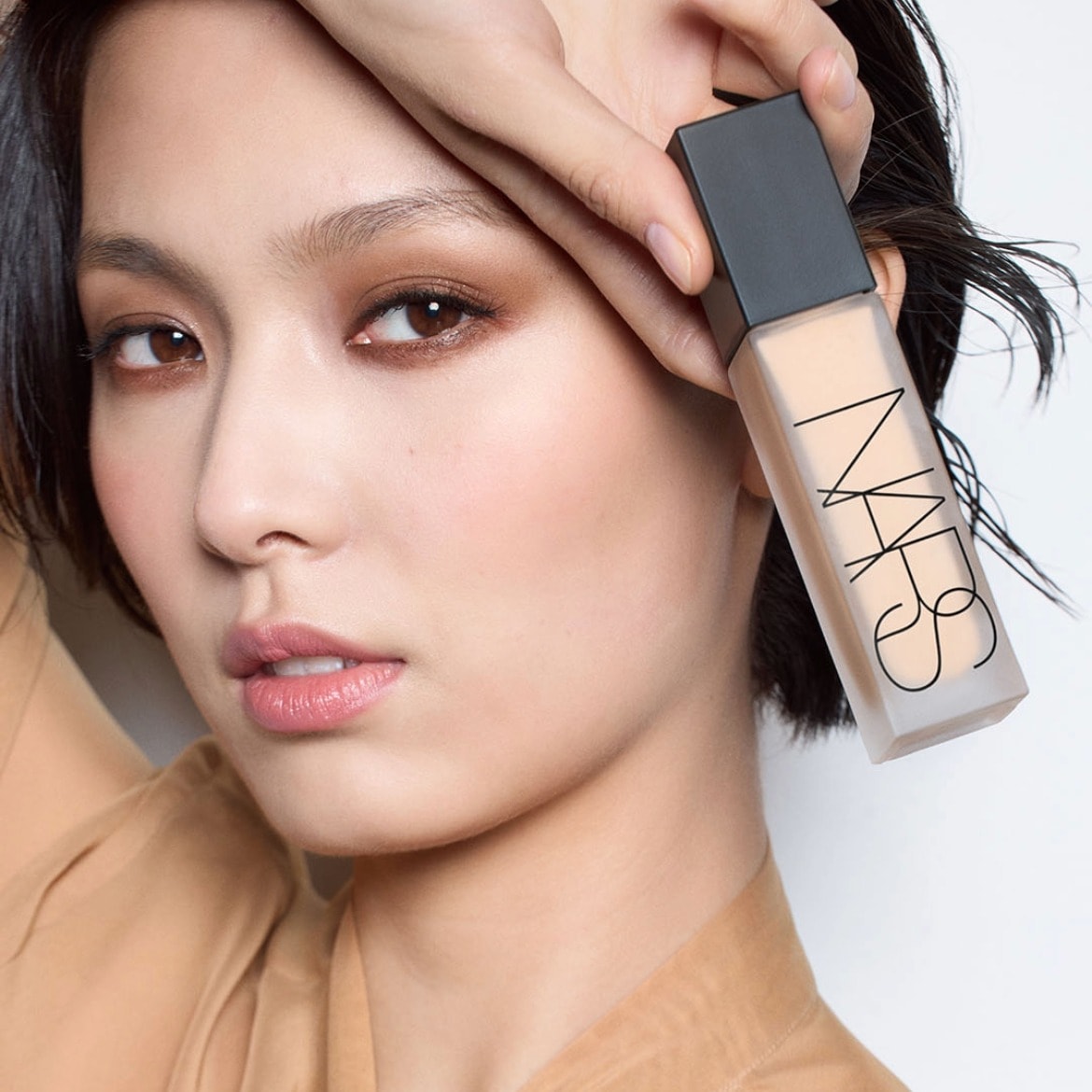 Een vrouw houdt een NARS foundationfles vast, met een matte dop en een doorzichtige fles gevuld met beige foundation, perfect passend bij haar huid.