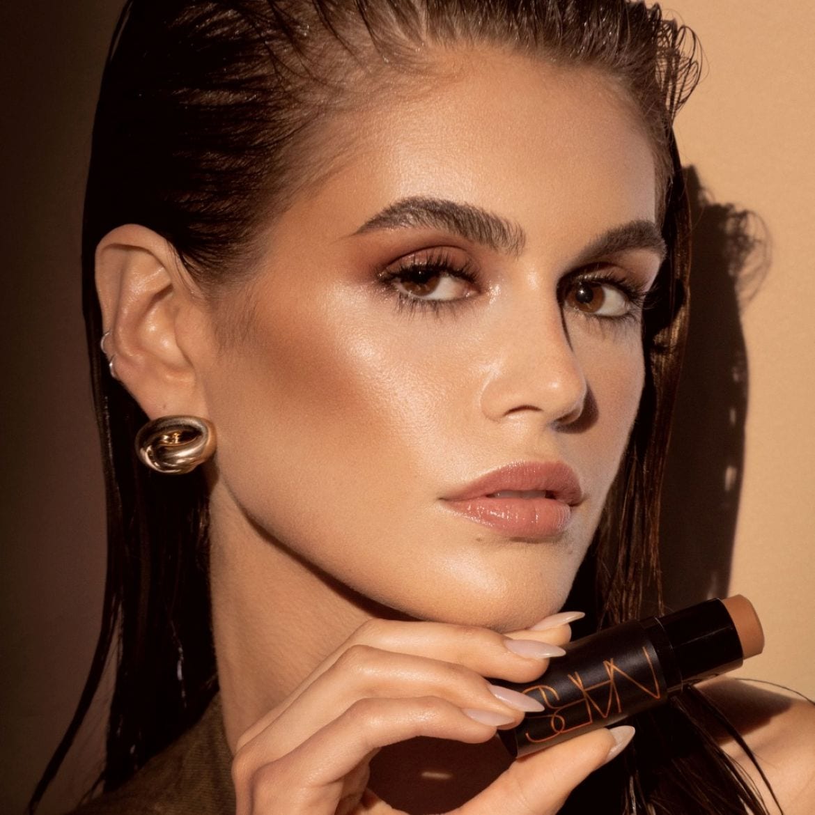Een vrouw met een stralende make-up houdt een NARS-contourstick vast, haar look afgemaakt met gouden oorbellen.
