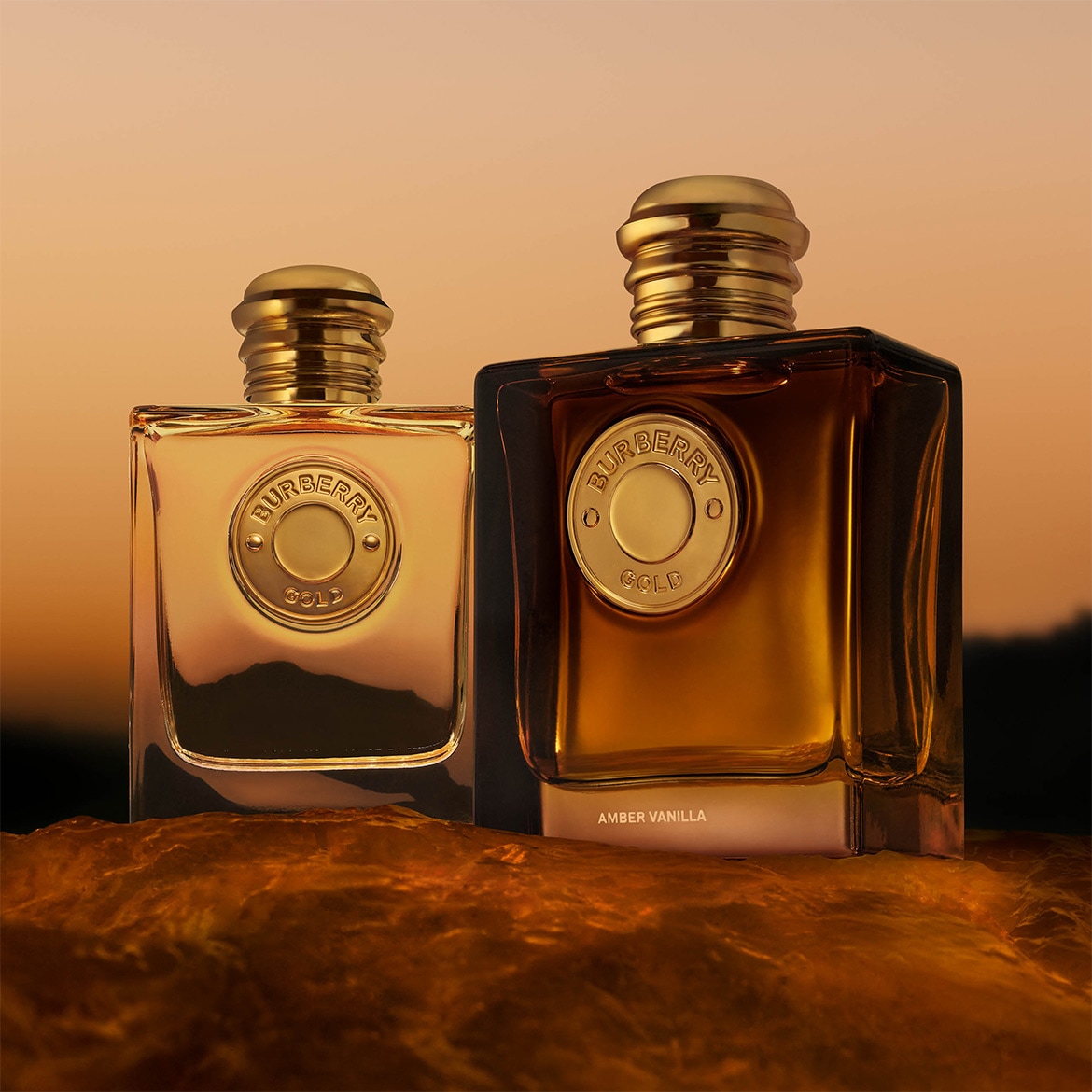 Twee flessen Burberry Gold parfum, de linker is lichtgoud en de rechter is donker amberkleurig, beide met een gouden dop en een gouden medaillon met het Burberry-logo, staand op een glanzend, roestbruin oppervlak tegen een oranje zonsondergang.