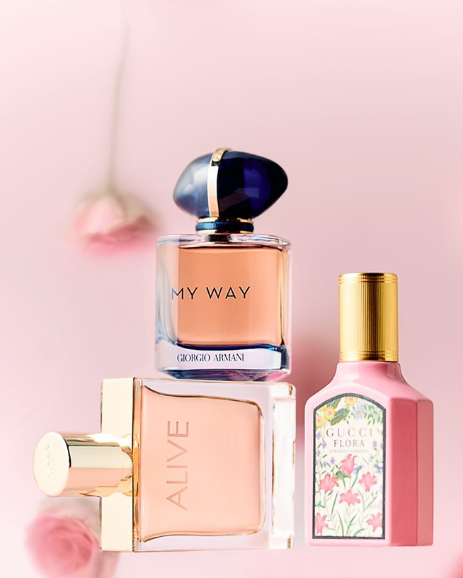 Ontdek een trio van parfums tegen een roze achtergrond: Giorgio Armani My Way, een vierkante fles met een blauwe dop, naast Boss Alive en Gucci Flora met bloemmotieven.