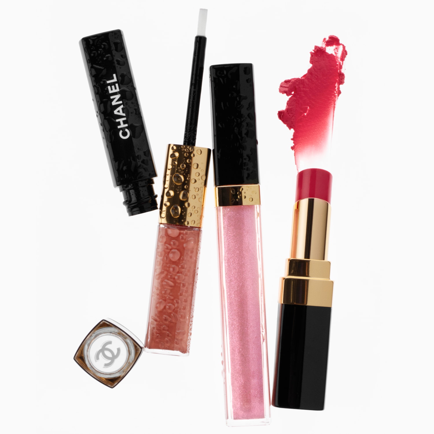 Een close-up van drie Chanel-lipglosses en een lippenstift, met waterdruppels op de verpakking, tegen een witte achtergrond.