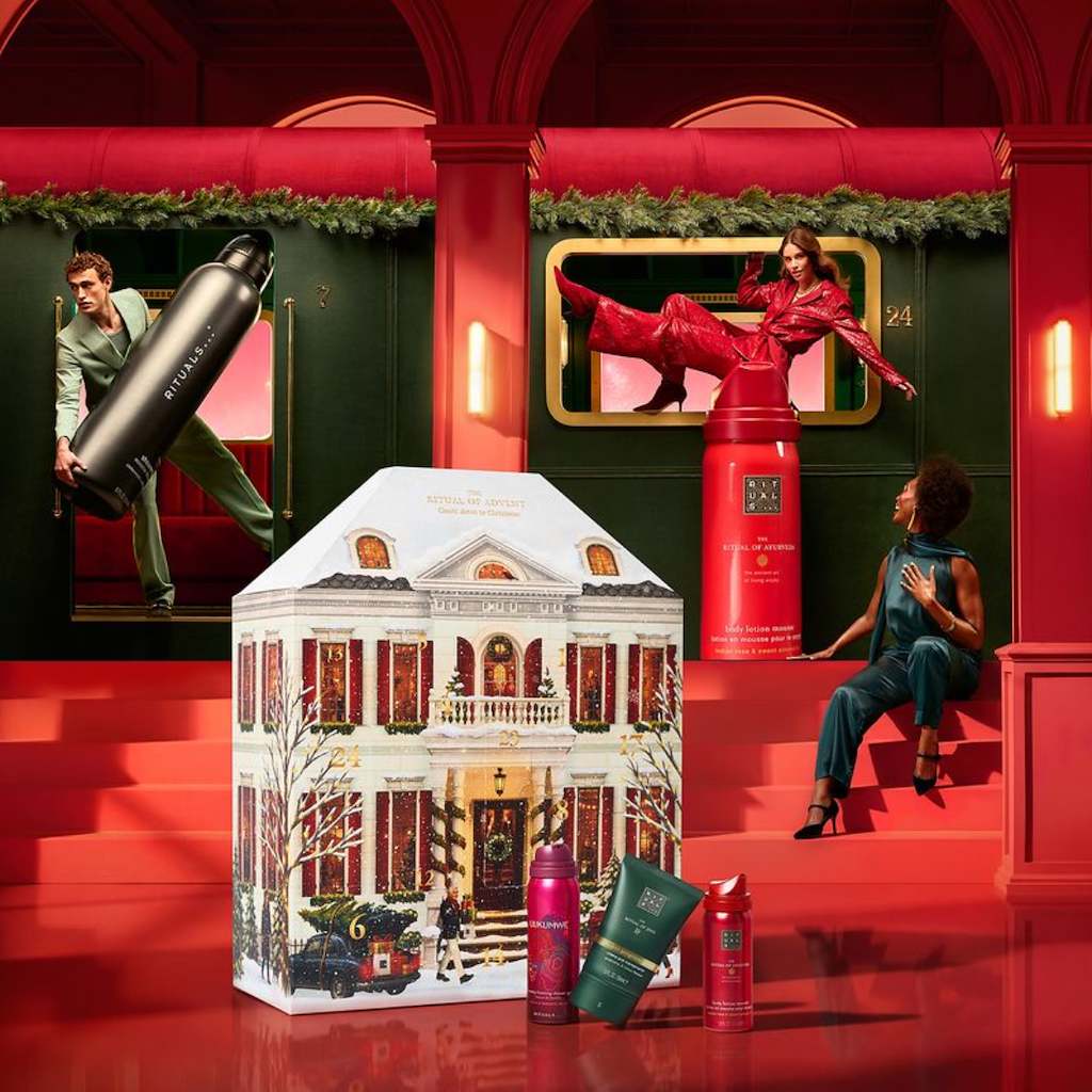 Een man in een groen pak stapt uit een treinwagon en draagt een grote zwarte fles van Rituals, met op de achtergrond een feestelijk versierd huis.